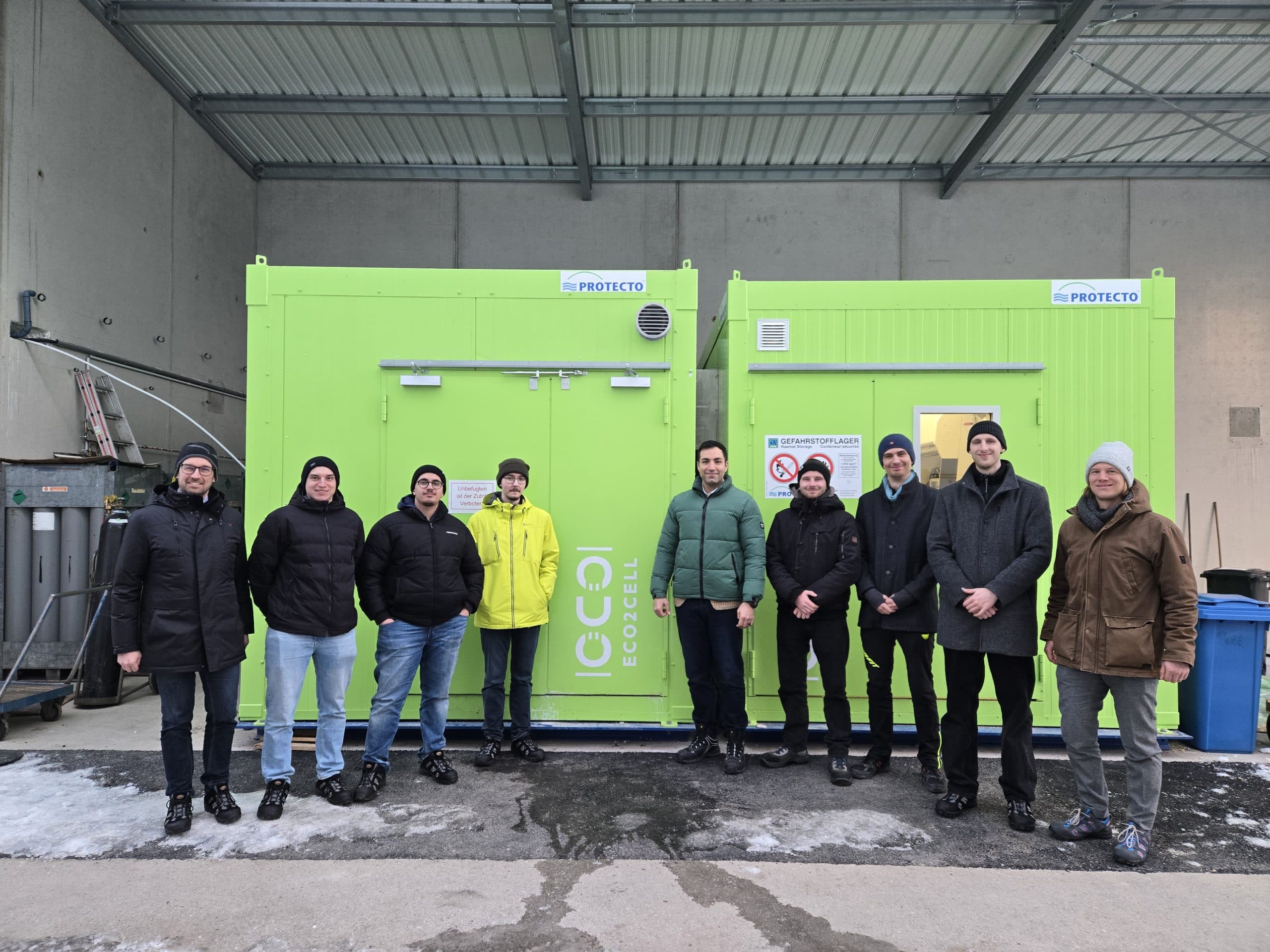 Neun Personen stehen in einer Reihe vor zwei hellgrünen Metallcontainern unter einem Unterstand, gekleidet in Winterkleidung. Der Boden ist teilweise mit Schnee bedeckt, und im Hintergrund sind Industriewände zu sehen.