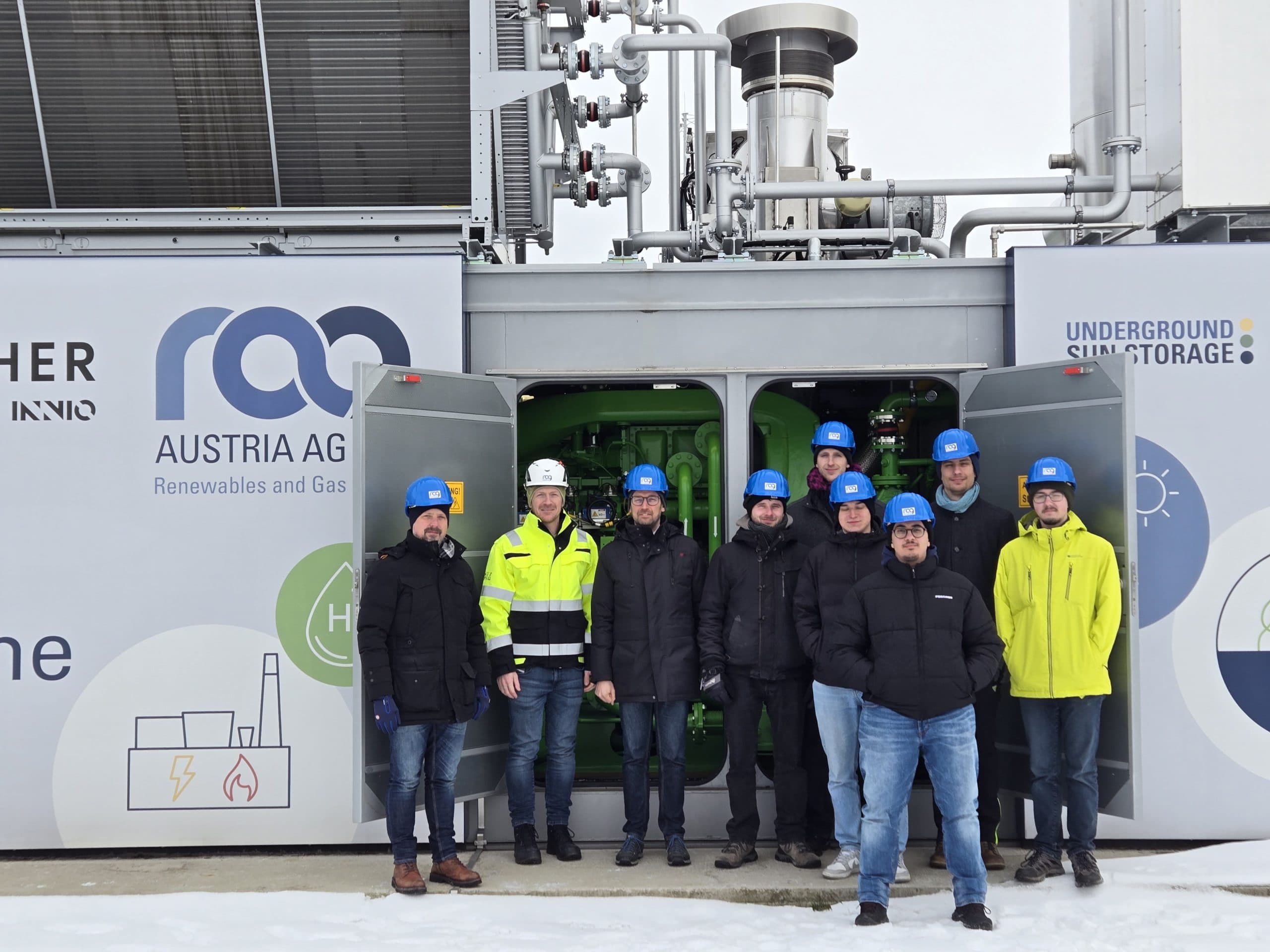 Eine Gruppe von neun Personen mit Schutzhelmen und Winterjacken steht vor einer Industrieanlage mit der Aufschrift AUSTRIA AG, hinter deren offenen Türen Geräte und Rohre sichtbar sind. Schnee bedeckt den Boden.