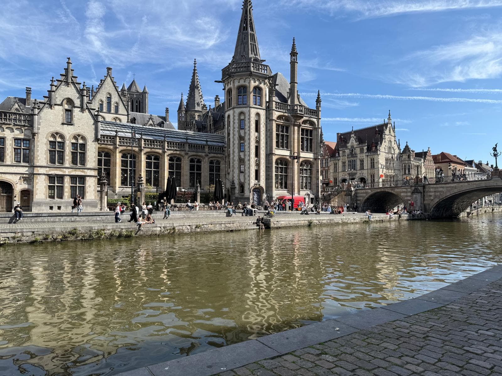 Historische Gebäude im gotischen Stil säumen einen Kanal in Gent, Belgien, und Menschen spazieren am Ufer entlang. Eine Steinbrücke überquert das Wasser unter einem strahlend blauen Himmel mit Wolkenfetzen.