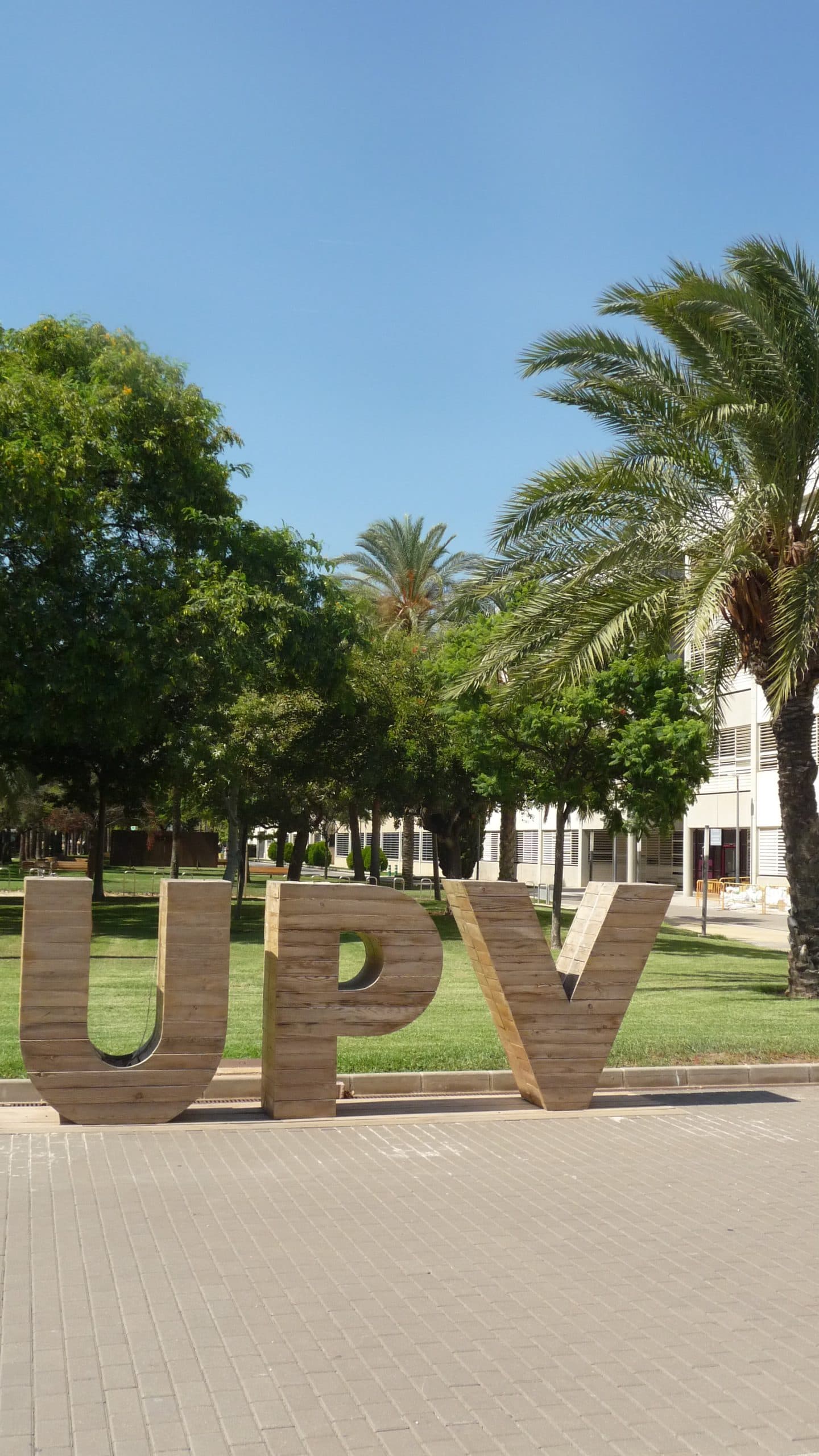 Große Holzbuchstaben mit dem Schriftzug UPV stehen im Freien auf einer gepflasterten Fläche, umgeben von grünen Bäumen und Palmen, mit Gebäuden im Hintergrund unter einem klaren blauen Himmel.