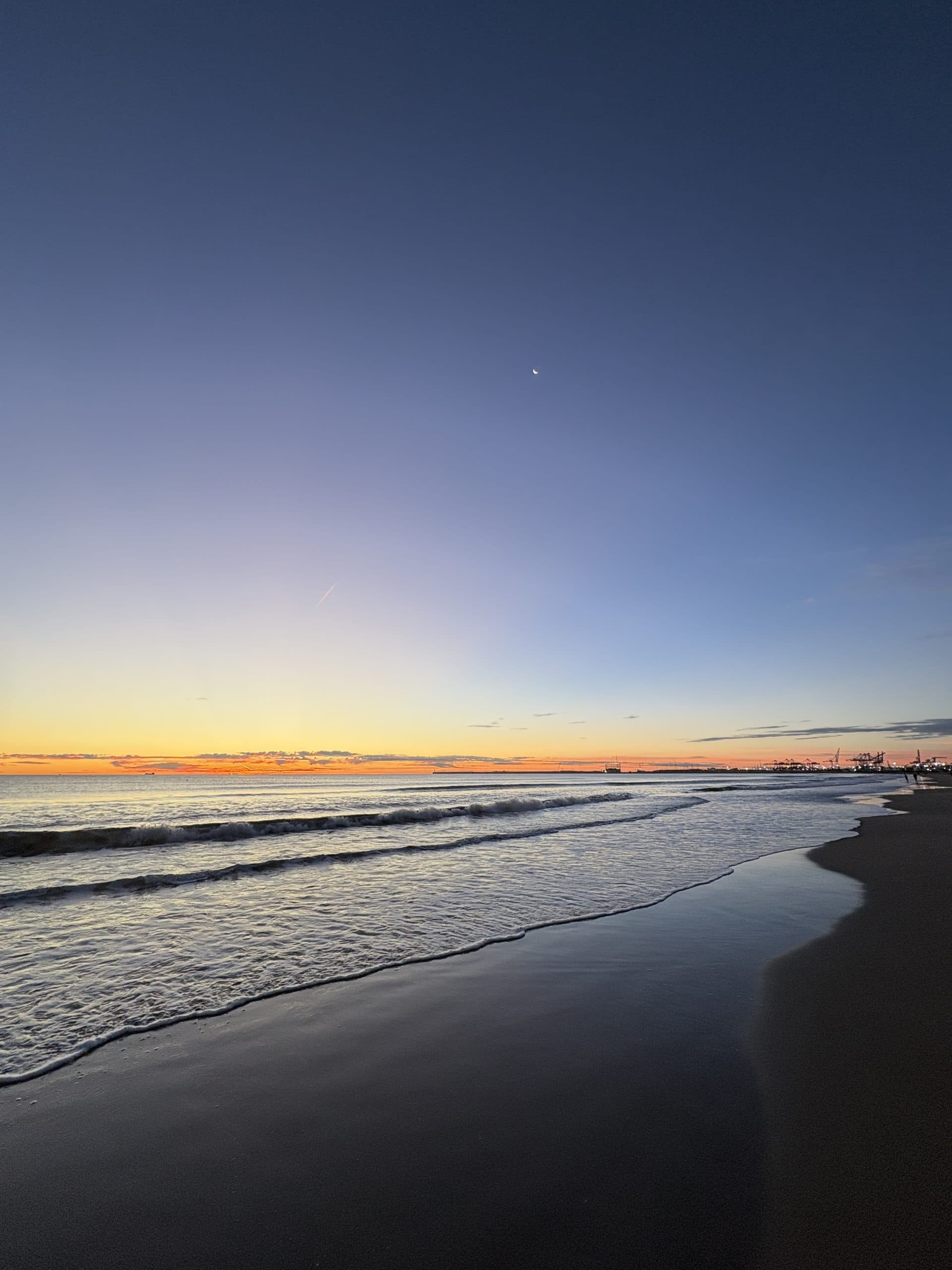 Ein ruhiger Strand bei Sonnenuntergang mit sanften Wellen, einem leuchtenden Horizont und einer kleinen Mondsichel an einem klaren, blauen Himmel. In der Ferne sind entlang der Küste Stadtsilhouetten zu erkennen.