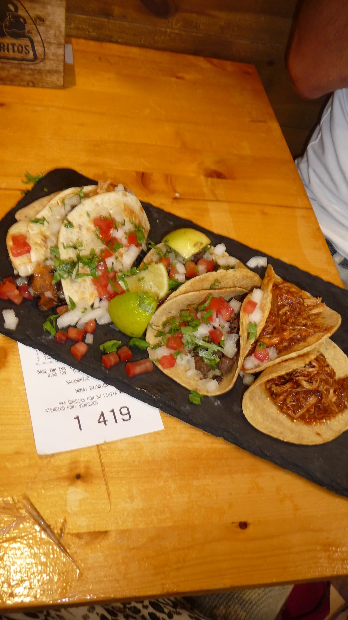 Ein schwarzer rechteckiger Teller mit vier Tacos, belegt mit gehackten Zwiebeln, Koriander und gewürfelten Tomaten, serviert mit Limettenspalten, auf einem Holztisch neben einer Restaurantrechnung.