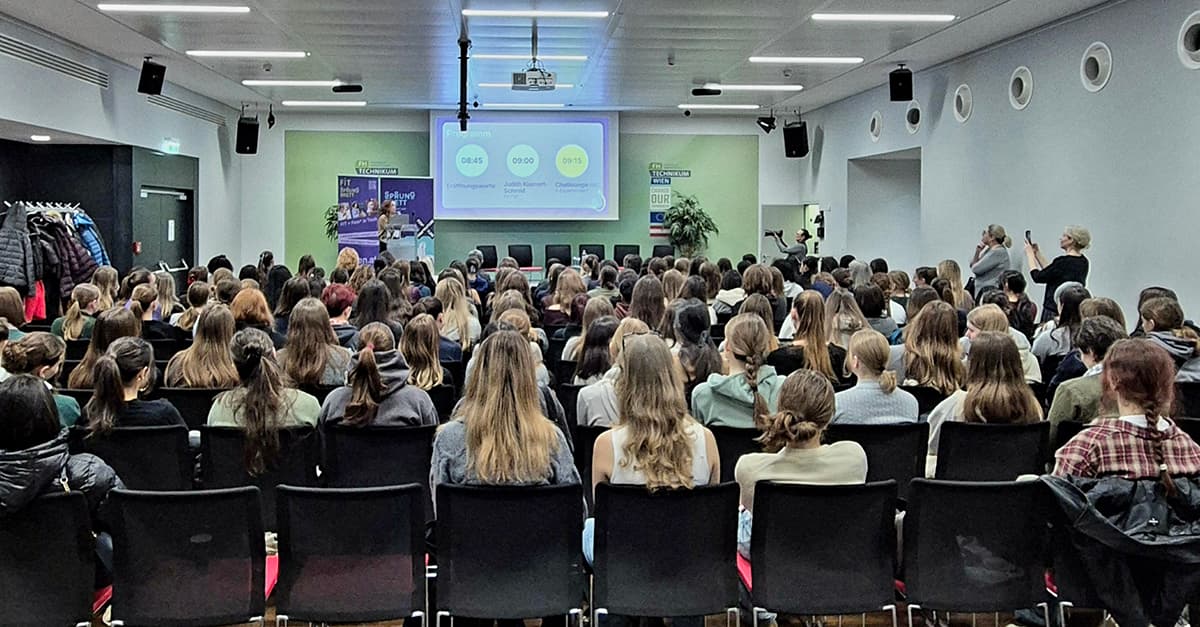 Eine große Gruppe von Personen sitzt während einer Präsentation in Stuhlreihen vor einem Bildschirm und Lautsprechern im vorderen Teil eines modernen Konferenzraums. Links hängen Mäntel an Regalen, und ein paar Leute stehen oder machen Fotos.