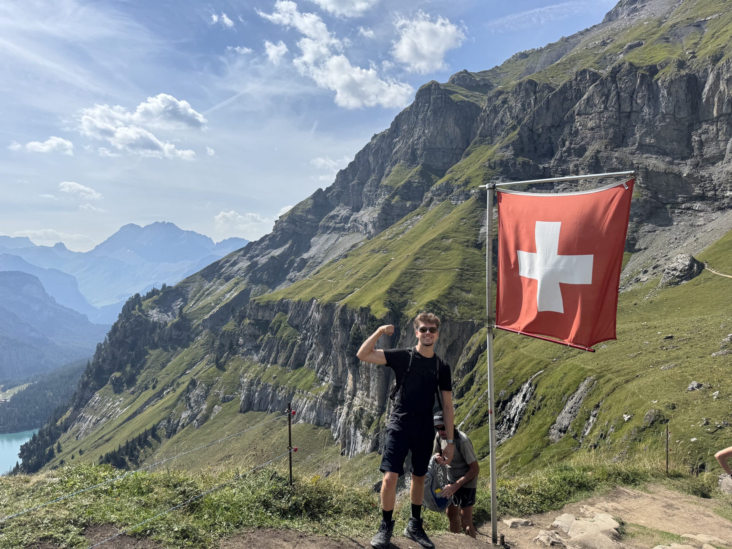 Ein Mann posiert mit gebeugtem Arm neben einer Schweizer Flagge auf einem Bergpfad, mit dramatischen grünen Hängen und schroffen Gipfeln im Hintergrund unter einem teilweise bewölkten Himmel.