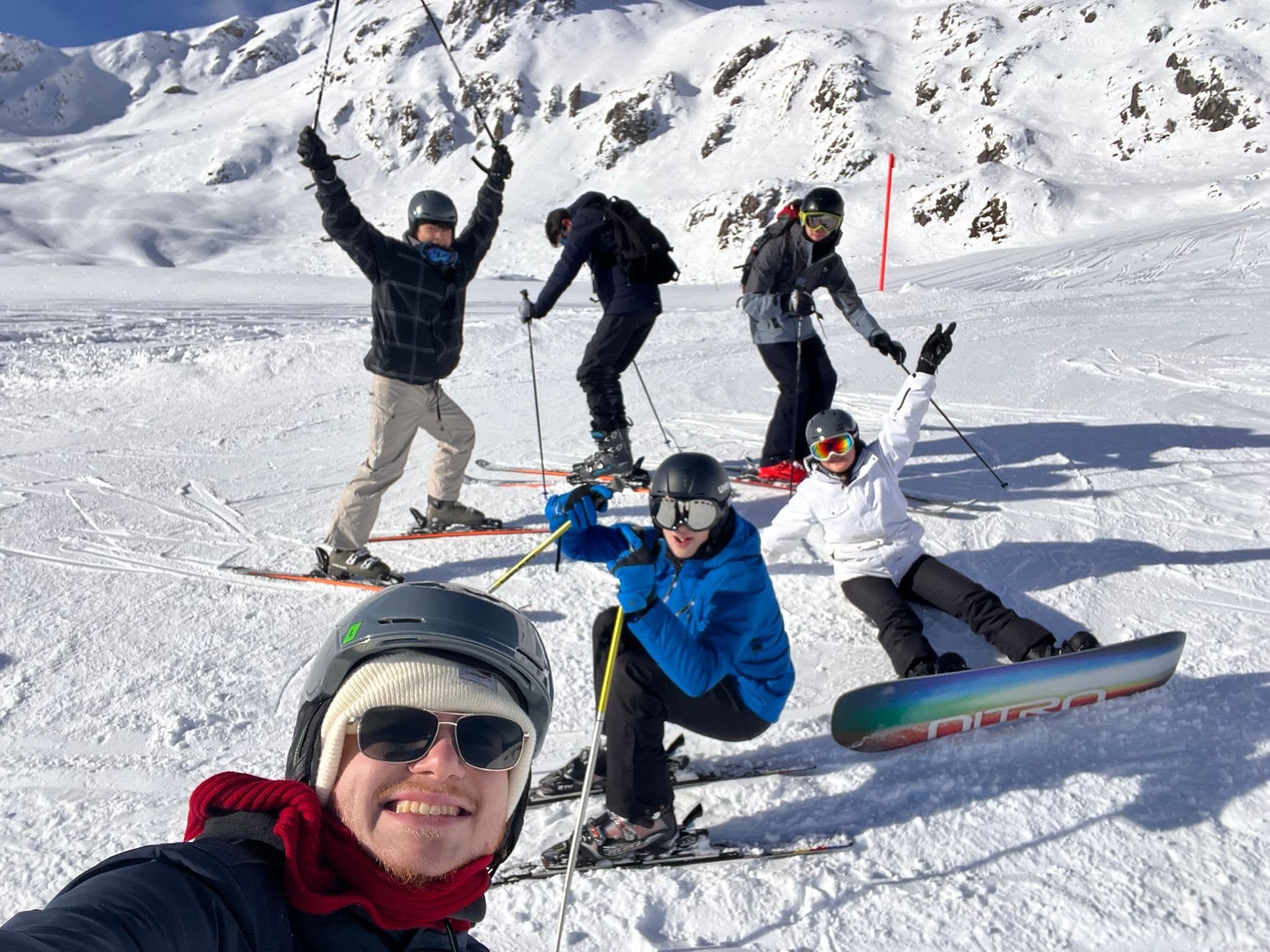 Eine Gruppe von sechs Personen in Skiausrüstung posiert und lächelt auf einem verschneiten Berghang; einige stehen auf Skiern, einer sitzt auf einem Snowboard, und die Sonne scheint auf die Berge hinter ihnen.