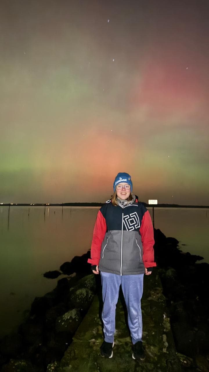 Eine Person, die eine blaue Mütze, eine Brille und eine rot-graue Jacke trägt, steht auf einem felsigen Pfad an einem ruhigen See in der Nacht, während bunte Polarlichter und Sterne am Himmel zu sehen sind.