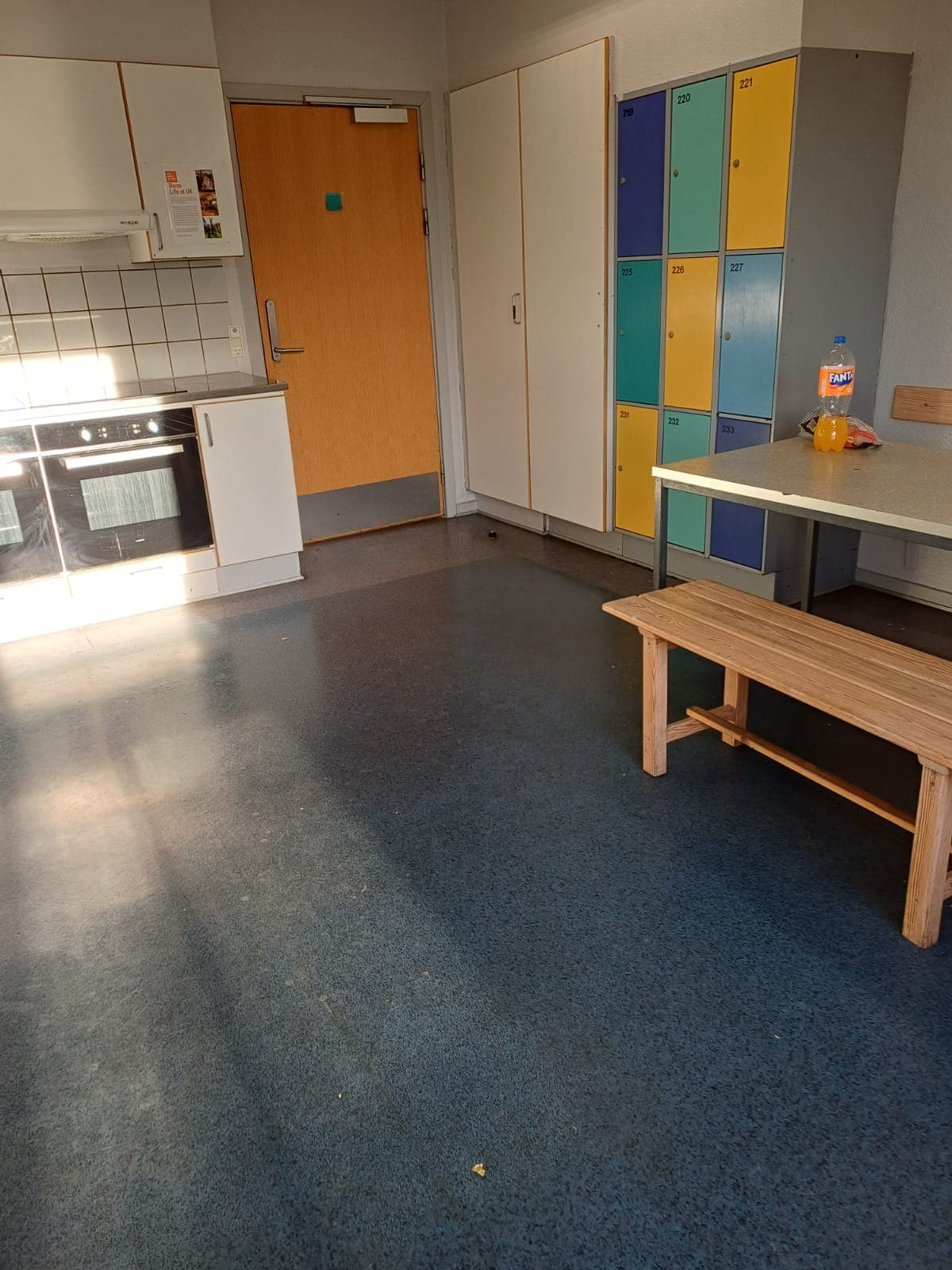 Eine Küche mit blauem Boden, einer Holzbank, einem Tisch, einem Ofen, weißen Schränken und bunten Schränken. Eine Plastikflasche und einige Gegenstände stehen auf dem Tisch, und Licht fällt durch ein Fenster.