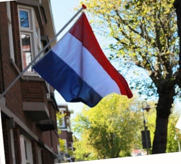 Eine niederländische Flagge mit horizontalen roten, weißen und blauen Streifen hängt an einem sonnigen Tag an einem Gebäude, mit Bäumen und einem klaren blauen Himmel im Hintergrund.