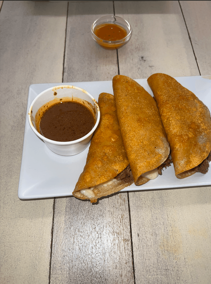 Zwei knusprige, mit Rindfleisch und geschmolzenem Käse gefüllte Birria-Tacos werden auf einem weißen Teller mit einer Schale dunkler, rotbrauner Consomé serviert. Eine weitere kleine Schale mit Consomé steht im Hintergrund auf einem Holztisch.