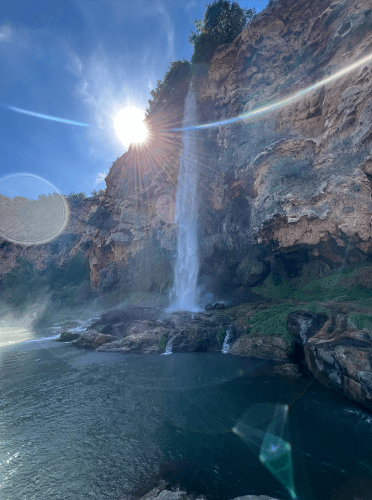 Ein hoher Wasserfall stürzt über eine Felsklippe in ein Becken, während das Sonnenlicht durch die Bäume über ihm fällt und Linsenreflexe und Nebel am strahlend blauen Himmel erzeugt.