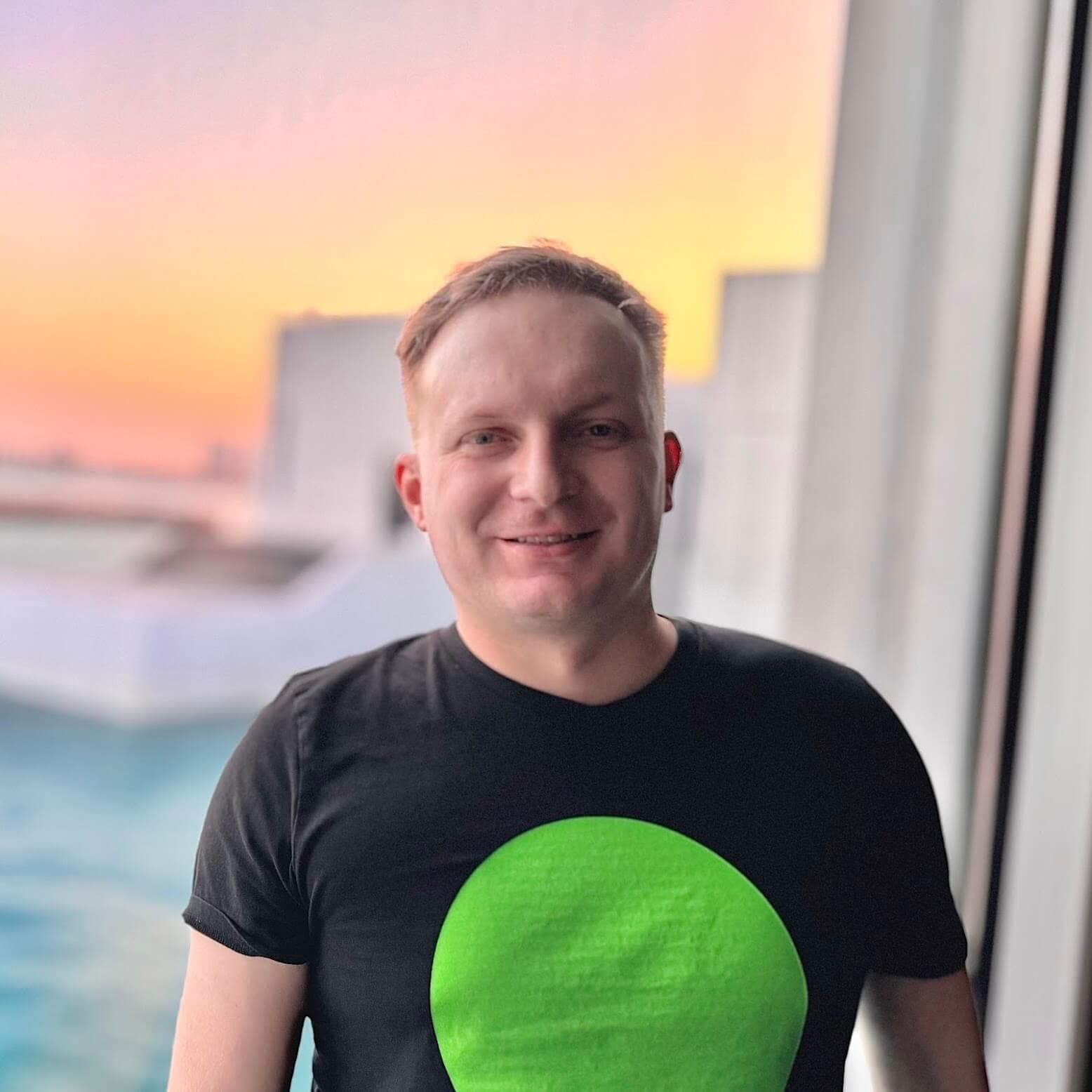 Eine Person mit kurzen Haaren lächelt, während sie drinnen an einem Fenster mit Blick auf das Wasser bei Sonnenuntergang steht. Sie trägt ein schwarzes T-Shirt mit einem großen grünen Kreis auf der Vorderseite. Draußen sind moderne weiße Gebäude zu sehen.