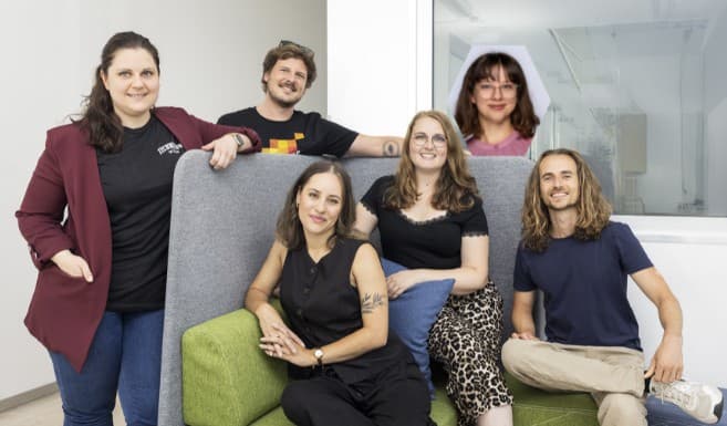 Sechs Personen posieren gemeinsam in einem modernen Büro; fünf von ihnen sitzen oder stehen um eine Couch herum, während eine Frau durch ein Glasfenster im Hintergrund lächelt. Sie wirken alle glücklich und entspannt.