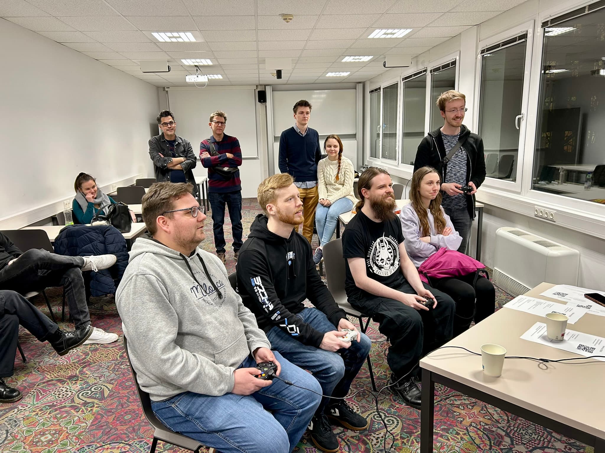 Eine Gruppe von Menschen ist in einem hellen Raum versammelt; vier von ihnen sitzen vorne und spielen ein Videospiel mit Controllern, während andere hinter ihnen stehen oder sitzen und lächeln. Auf dem Tisch daneben liegen Papiere und Tassen.