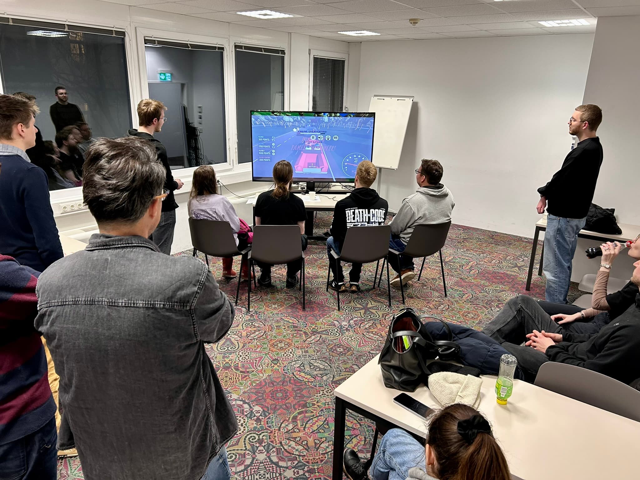 Eine Gruppe von Menschen in einer zwanglosen Innenumgebung beobachtet zwei Spieler, die in einem Videospiel gegeneinander antreten, das auf einem großen Bildschirm angezeigt wird. Einige sitzen, andere stehen herum, und im Hintergrund ist ein Flipchart zu sehen.