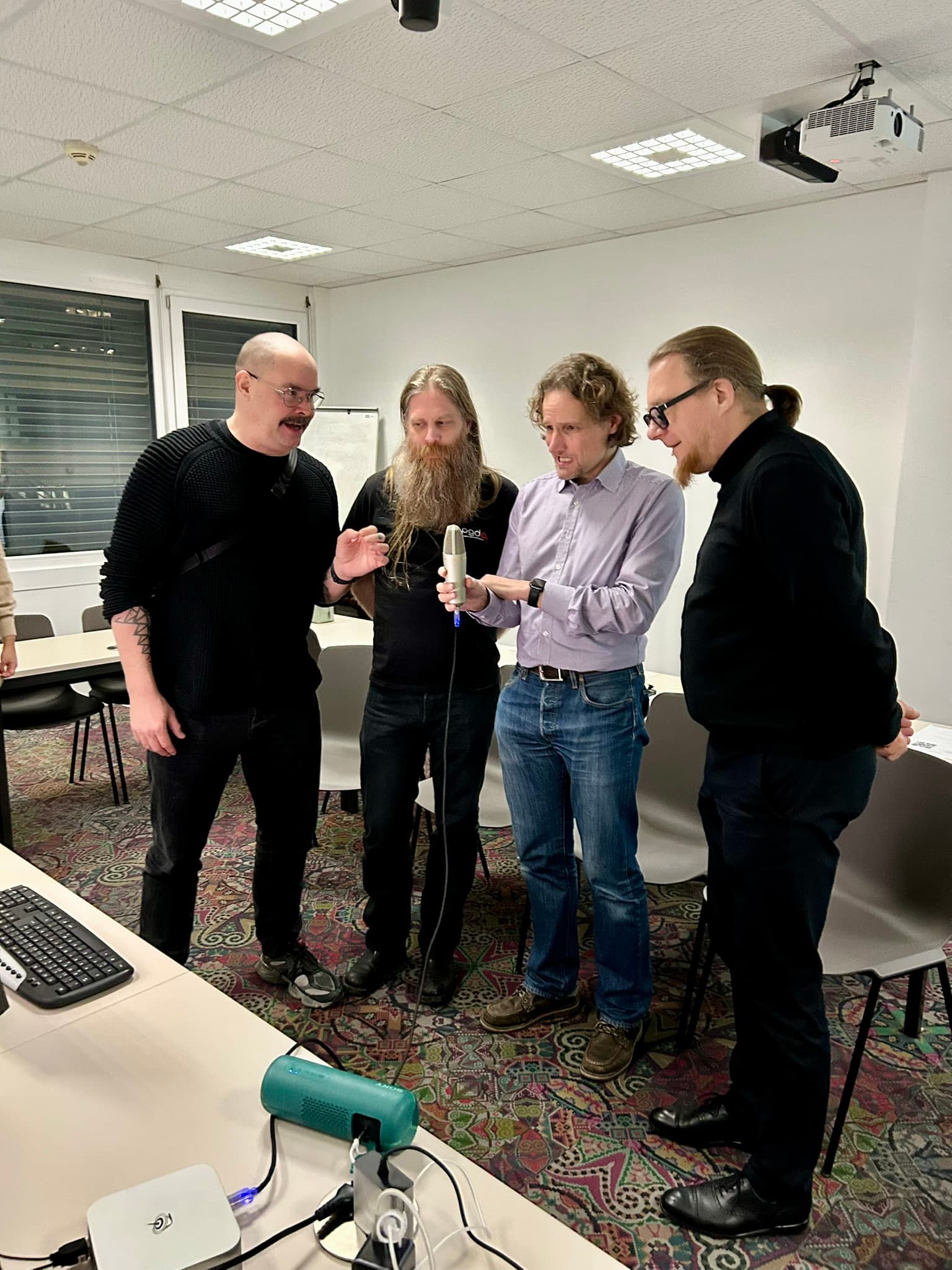 Vier Männer stehen zusammen in einem Büro und untersuchen ein Mikrofon, das einer von ihnen in der Hand hält. Sie scheinen eine Diskussion zu führen. In dem Raum sind Tische, Stühle, eine Computertastatur und ein Projektor zu sehen.