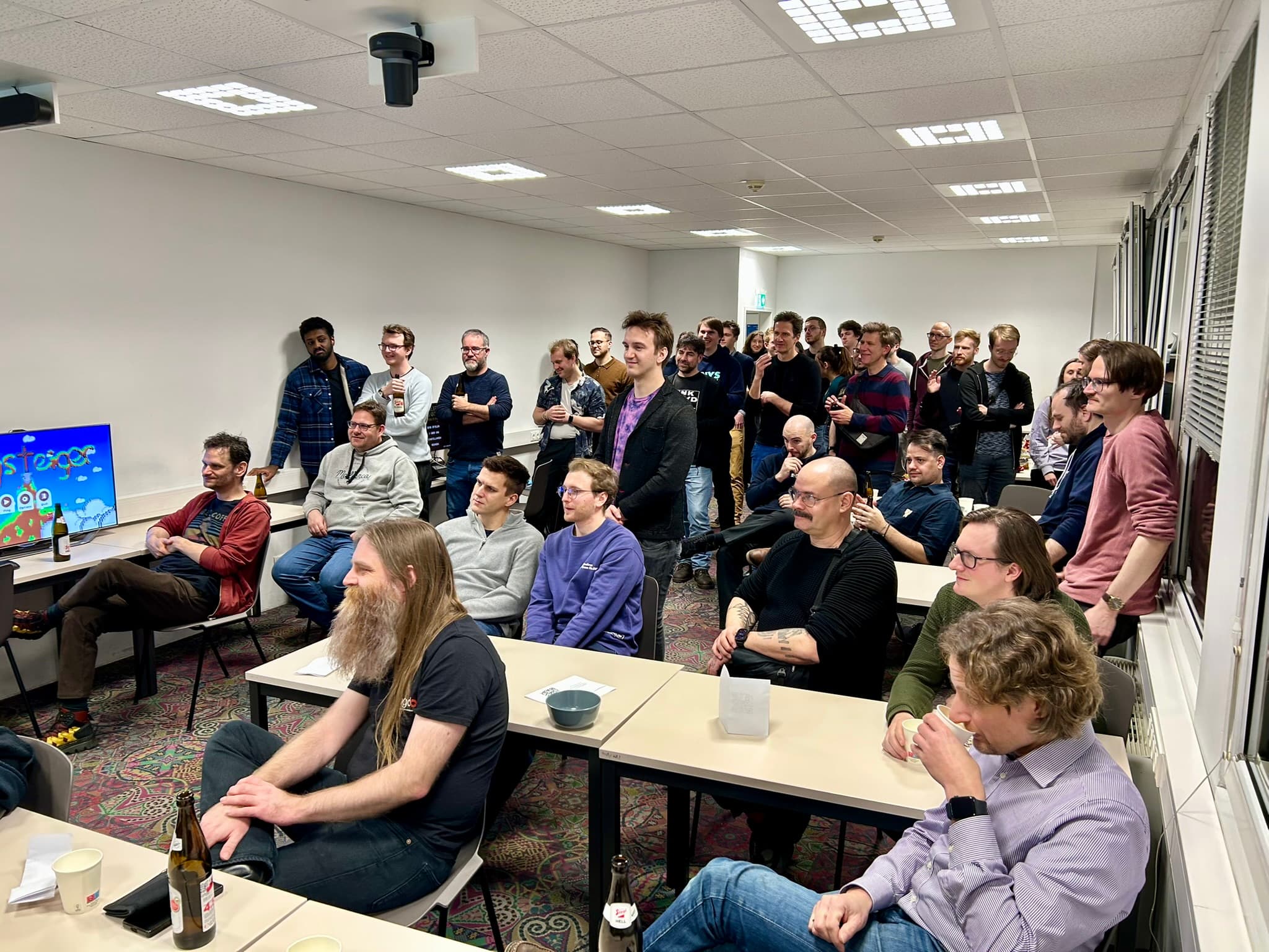 Eine Gruppe von Menschen, meist Männer, ist in einem hell erleuchteten Raum versammelt, sitzt und steht um Tische herum und verfolgt aufmerksam eine Präsentation oder ein Spiel auf einem Bildschirm im vorderen Teil des Raums. Einige lächeln und trinken.