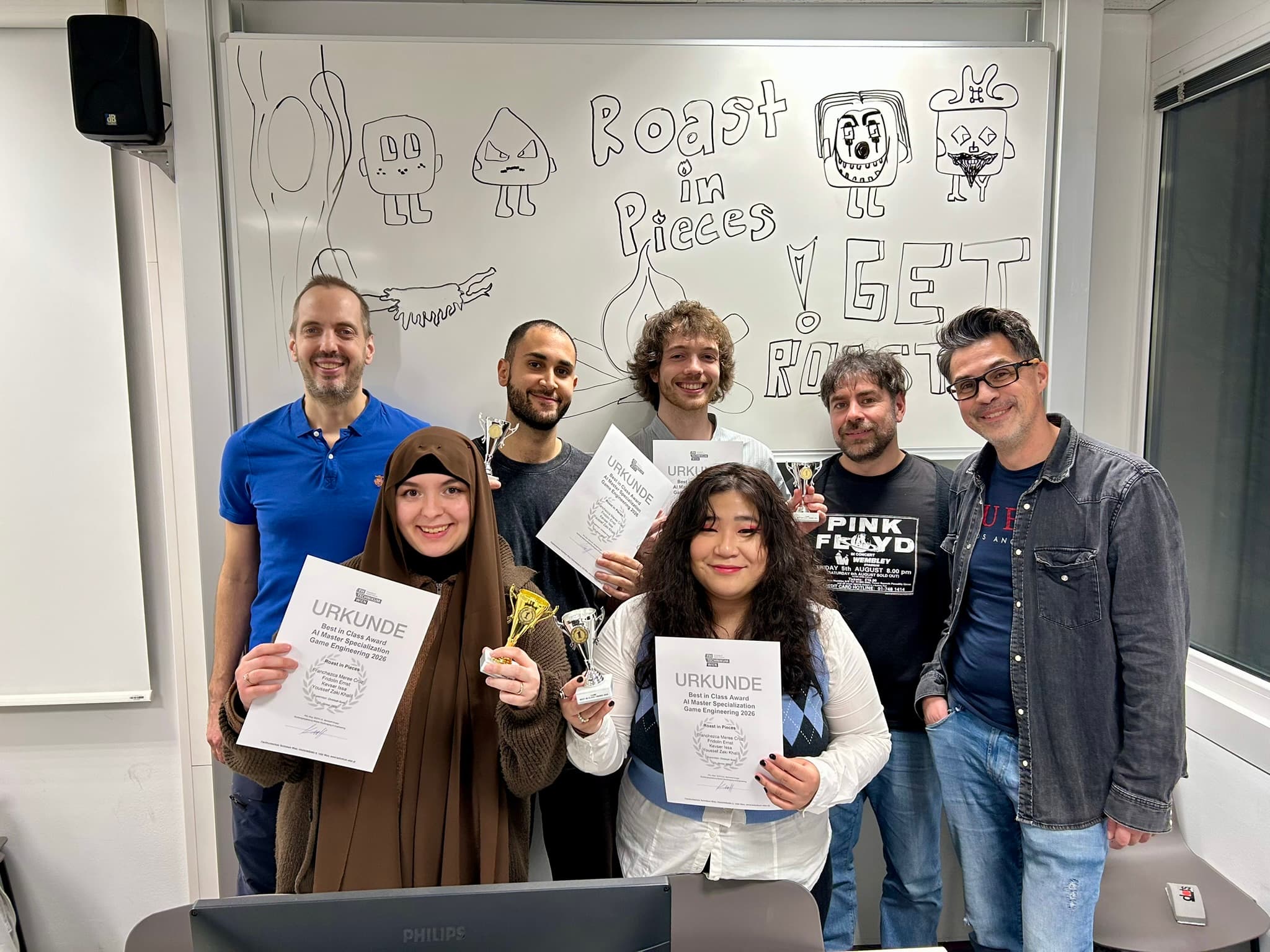 Sieben Personen stehen lächelnd in einem Klassenzimmer und halten Urkunden und kleine Trophäen in der Hand. Hinter ihnen steht eine Tafel mit Cartoon-Zeichnungen und der Aufschrift 'Roast in Pieces' und 'GET ROASTED'.