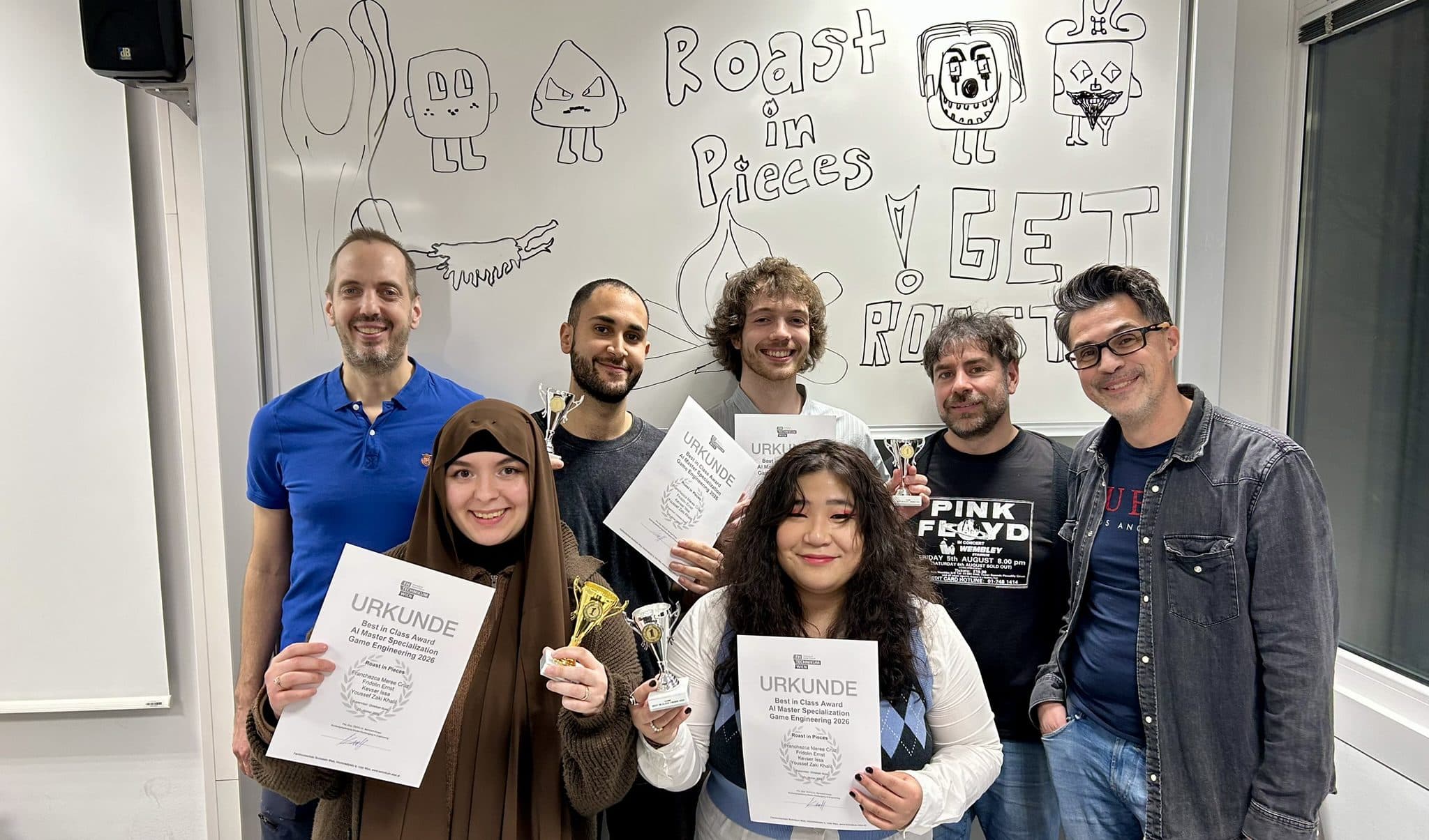 Sechs Personen stehen in einem Klassenzimmer, einige halten Urkunden und kleine Trophäen in der Hand und lächeln in die Kamera. Hinter ihnen steht eine Tafel mit Cartoon-Zeichnungen und der Aufschrift "Roast in Pieces".