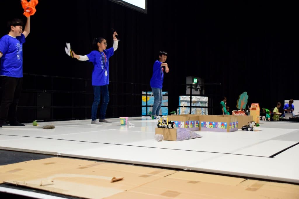 Drei junge Menschen in blauen Hemden stehen auf einer Bühne mit einem Roboterwettbewerb, der aus kleinen Robotern, Hindernissen und bunten Barrieren besteht. Eine Person hebt einen Gegenstand hoch, während die anderen sich auf die Roboter konzentrieren.