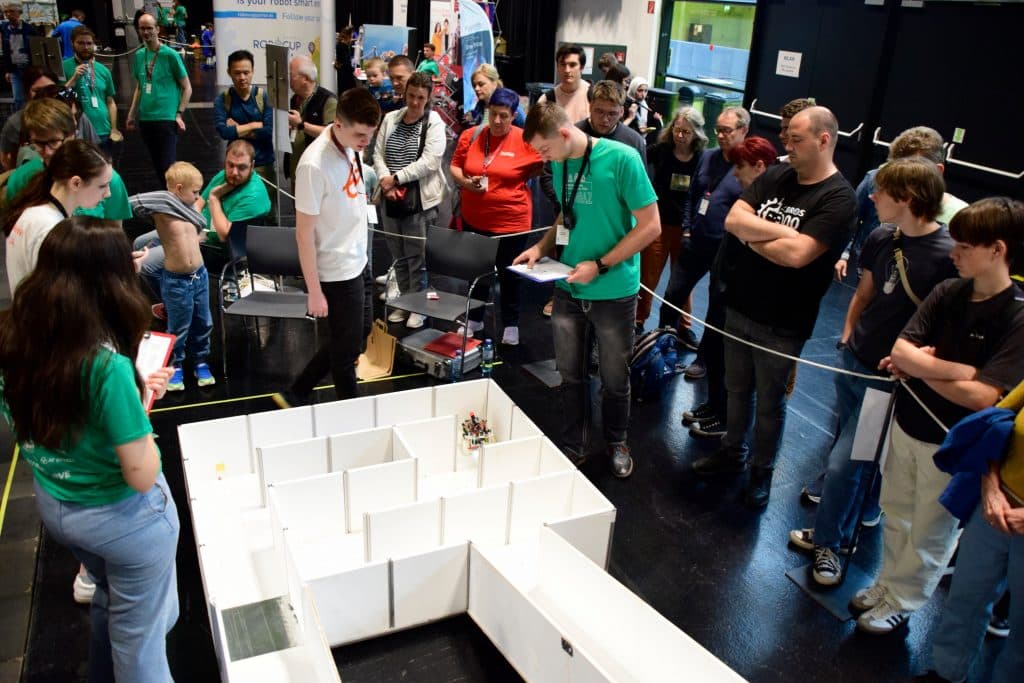 Eine Gruppe von Menschen steht um ein weißes Labyrinth auf schwarzem Boden und beobachtet, wie Teilnehmer einen kleinen Roboter durch das Labyrinth führen. Einige Personen halten Klemmbretter, was auf einen Wettbewerb oder eine Bewertung hinweist.