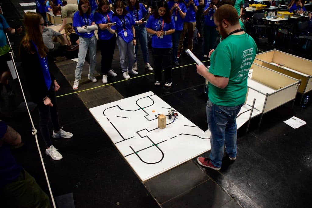 Eine Gruppe von Menschen beobachtet, wie ein Mann mit einem Klemmbrett einen kleinen Roboter beobachtet, der auf einer weißen Matte einen Parcours abfährt, wahrscheinlich während eines Robotikwettbewerbs oder einer Demonstration.