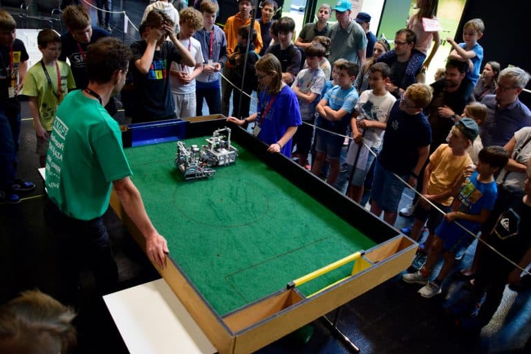 Eine Gruppe von Kindern und Erwachsenen versammelt sich um ein Tischfußballfeld und beobachtet zwei Robotik-Teams, die kleine Roboter in einem Spiel steuern. Die Atmosphäre ist lebhaft und konzentriert, während alle den Wettbewerb beobachten.