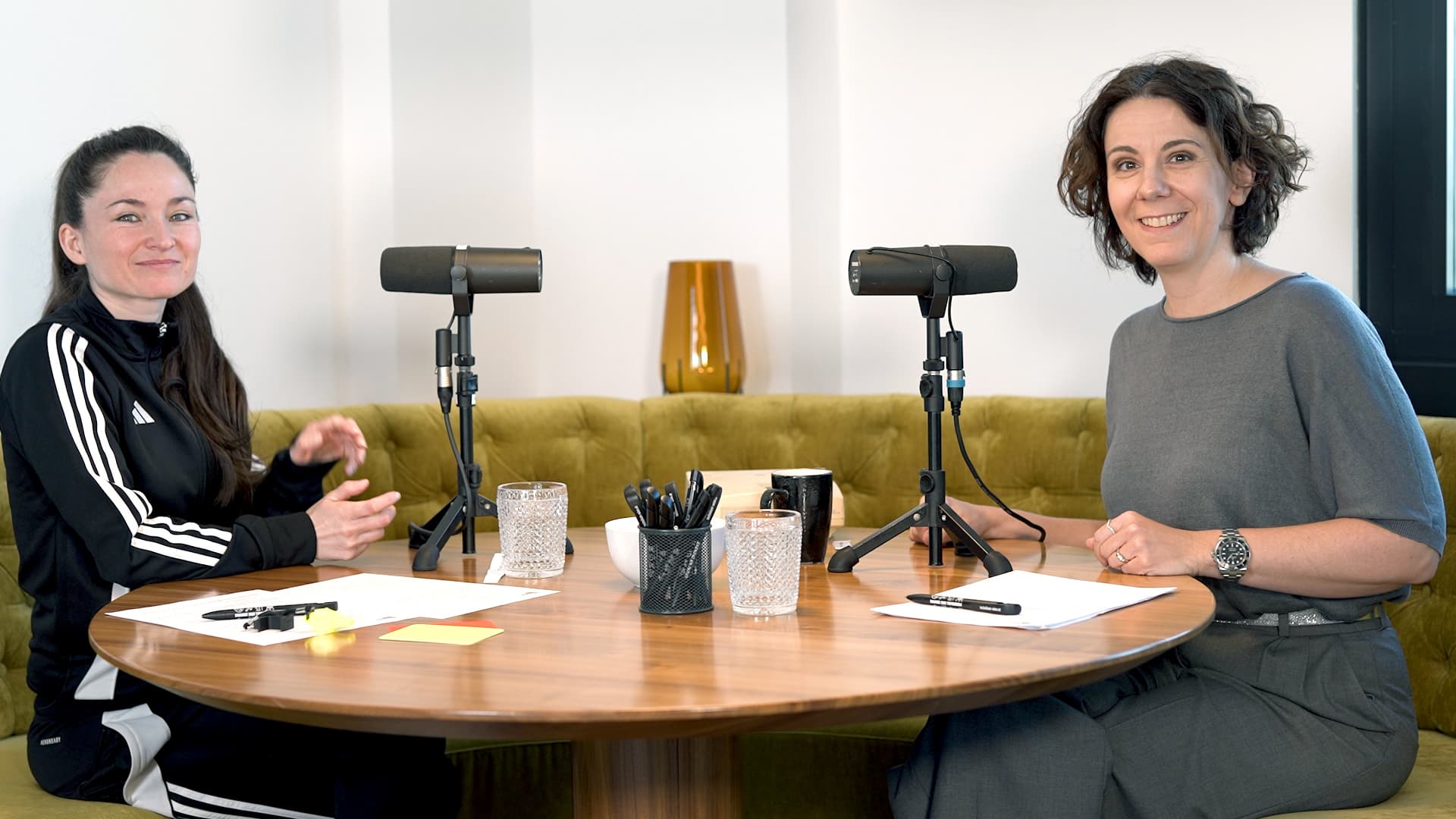 Zwei Frauen sitzen an einem runden Holztisch mit Mikrofonen, Papieren, Stiften und Gläsern mit Wasser und bereiten sich auf einen Podcast oder ein Interview vor. Sie lächeln in einem modernen, hell erleuchteten Raum mit einer grünen Couch hinter ihnen.