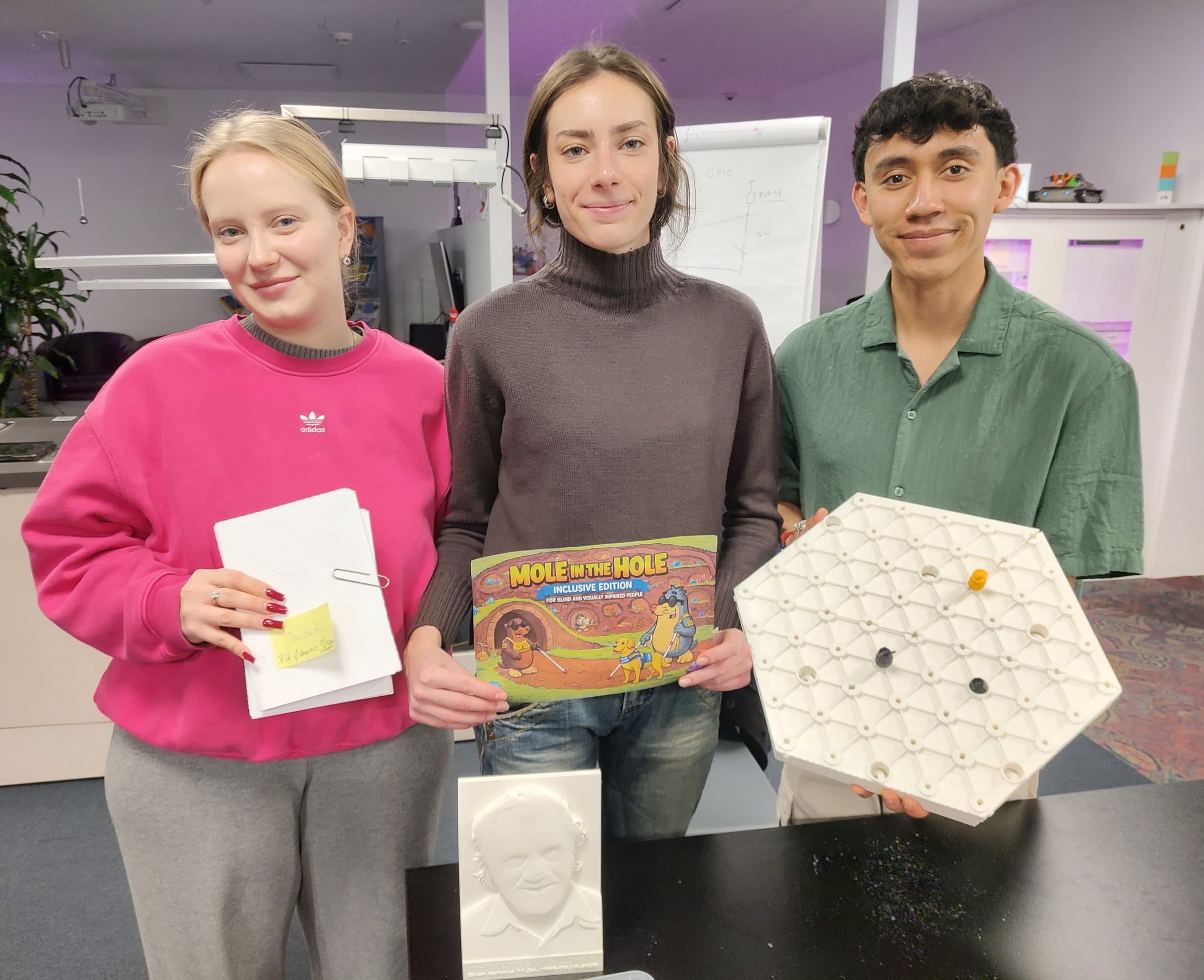 Student team holding the adapted board game "Mole in the Hole" and the rule book. On the table a 3D printed portrait of the co-designer Erich Schmid is standing.