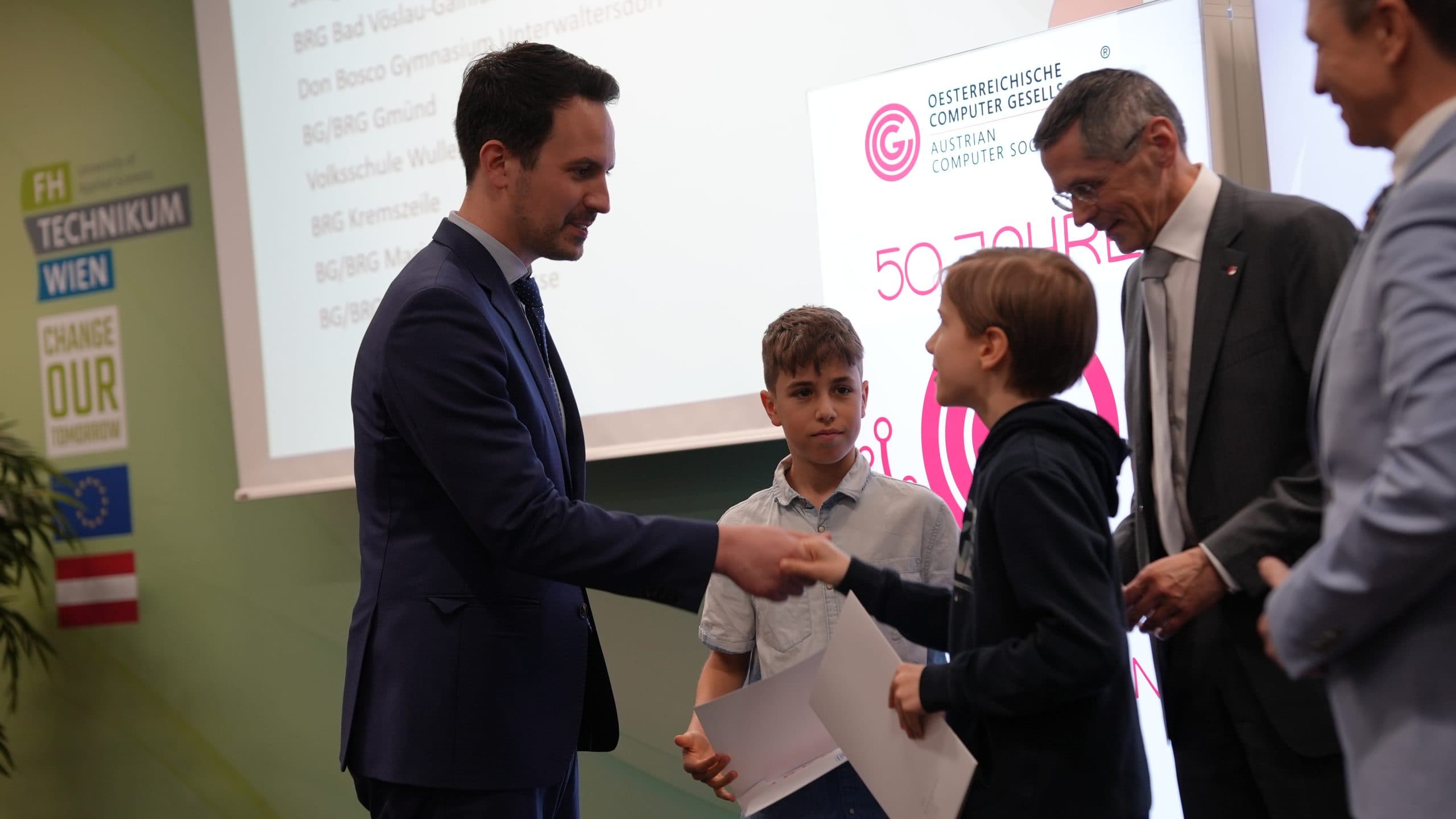 Ein Mann im Anzug schüttelt einem lächelnden Jungen, der eine Urkunde hält, die Hand, während ein anderer Junge und zwei Männer bei einer Preisverleihung zusehen. Im Hintergrund sind ein Präsentationsbildschirm und ein Veranstaltungsbanner zu sehen.