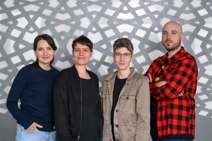 Vier Personen stehen nebeneinander vor einer gemusterten grauen und weißen Wand und lächeln oder schauen in die Kamera. Sie tragen Freizeitkleidung, darunter ein blaues Hemd, eine schwarze Jacke, eine hellbraune Jacke und ein rot kariertes Hemd.