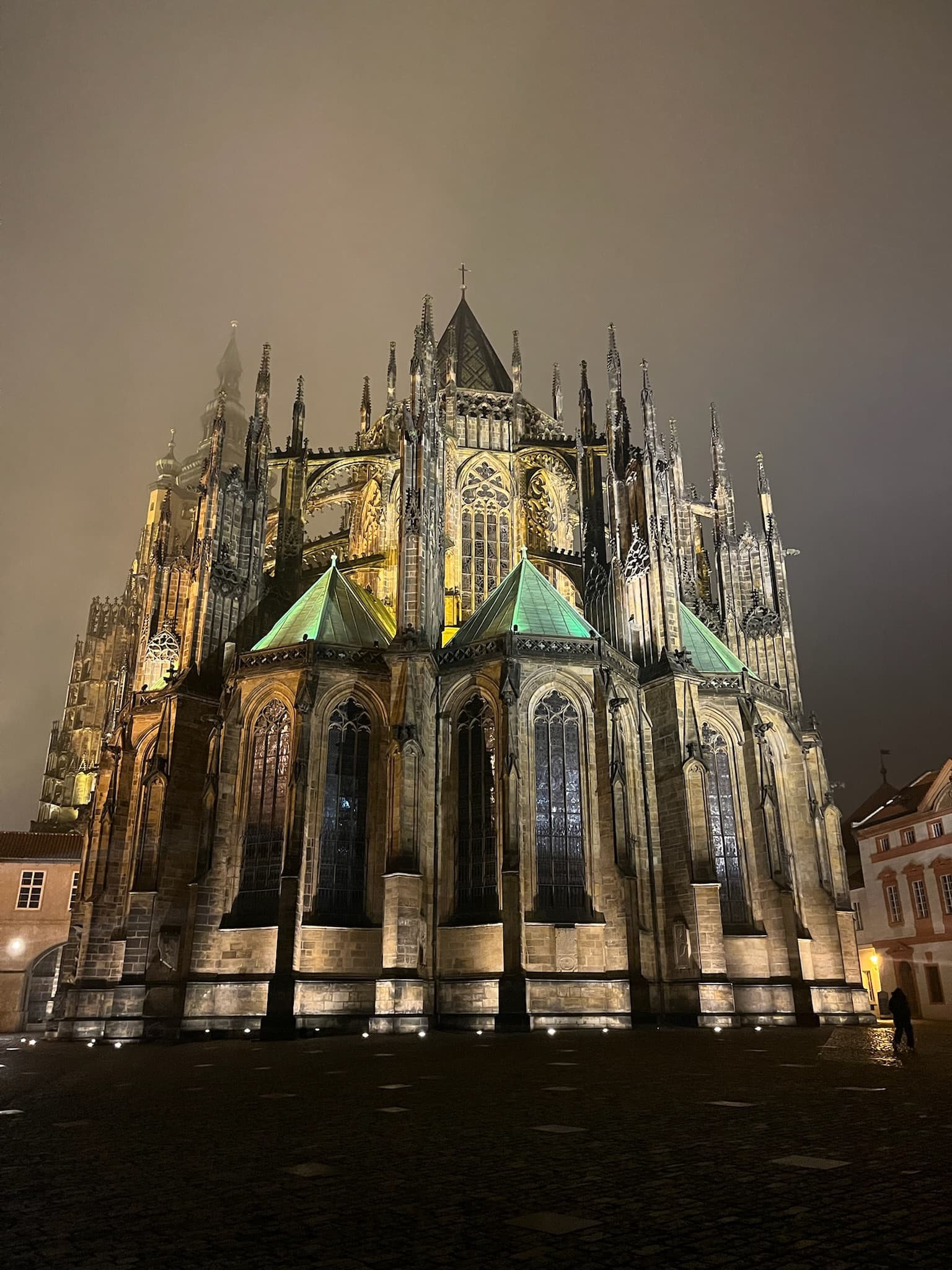 Eine gotische Kathedrale mit hohen Türmen und kunstvollen Details ist nachts dramatisch beleuchtet, umgeben von einem dunstigen, nebligen Himmel. Das steinerne Äußere des Gebäudes und die spitzen Bögen stechen hervor, und an der Basis leuchten Lichter.