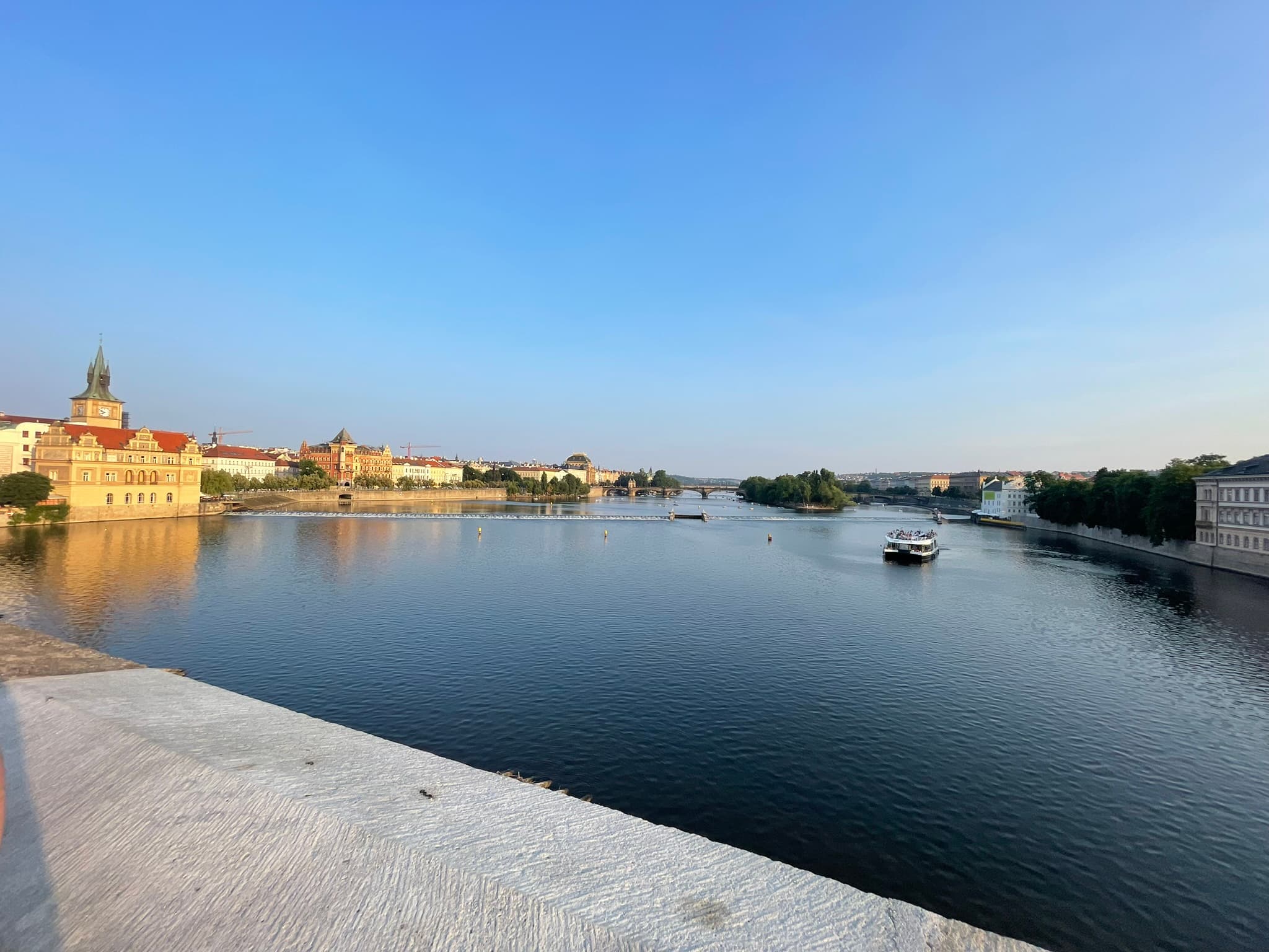 Ein breiter Fluss mit ruhigem Wasser fließt durch eine historische Stadt mit bunten Gebäuden und einem Kirchturm auf der linken Seite. Ein Boot ist auf dem Fluss, und der Himmel ist klar und blau.