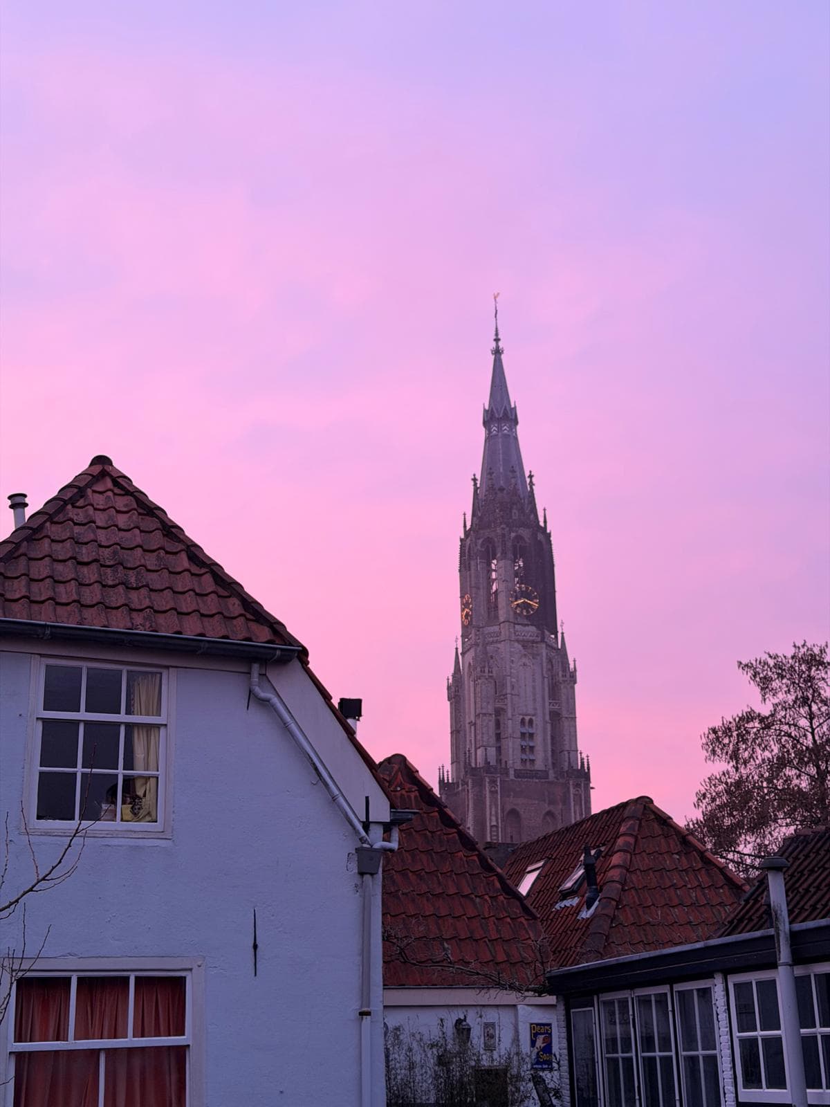 Ein hoher Kirchturm im gotischen Stil erhebt sich über den Dächern weißer Häuser mit roten Ziegeln vor einem leuchtend rosafarbenen und violetten Sonnenuntergangshimmel.