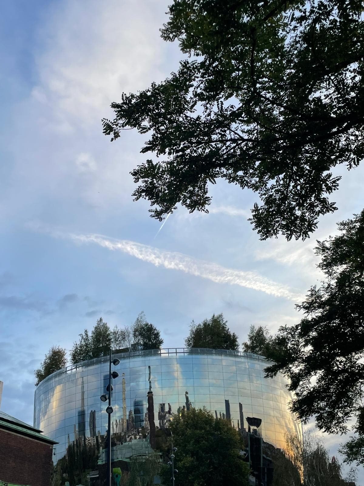 Ein modernes, rundes Glasgebäude mit Bäumen auf dem Dach spiegelt die Skyline der Stadt unter einem teilweise bewölkten Himmel wider. Üppige Baumzweige umrahmen die Szene in der oberen rechten Ecke.