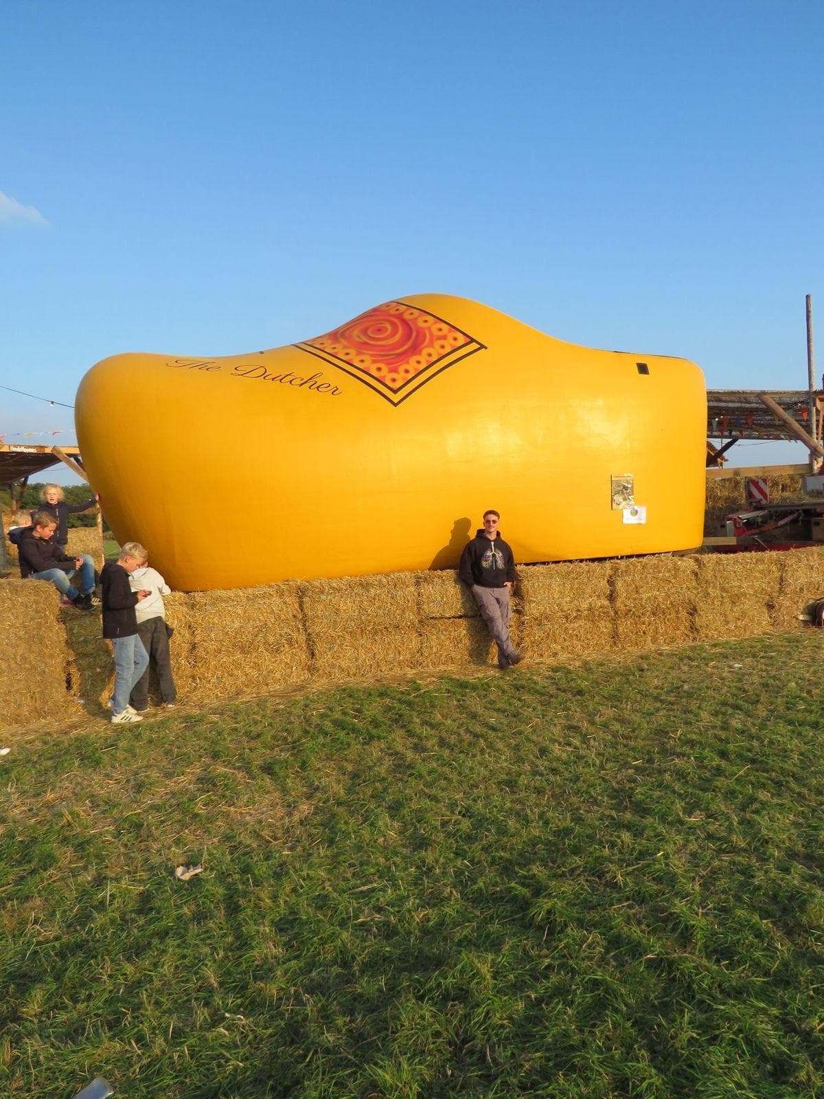 Eine riesige gelbe Holzpantoffel-Skulptur sitzt auf Heuballen im Freien, und drei Menschen stehen in der Nähe im Gras unter einem klaren blauen Himmel.