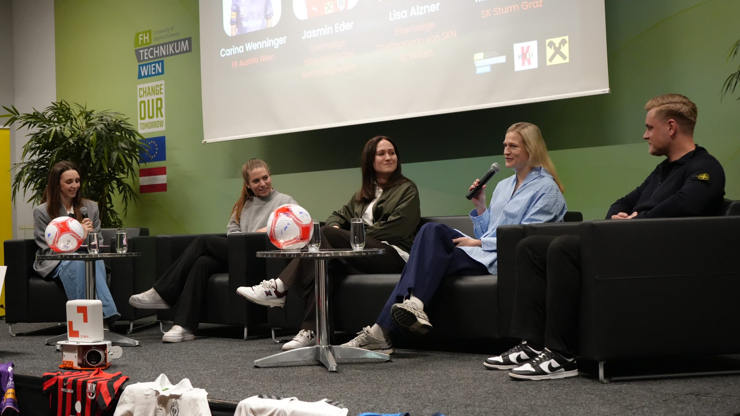 Fünf Personen sitzen in einer Podiumsdiskussion auf einer Bühne, mit Mikrofonen und Markenfußbällen auf dem Tisch. Im Hintergrund sind ein Präsentationsbildschirm und Grünflächen zu sehen.