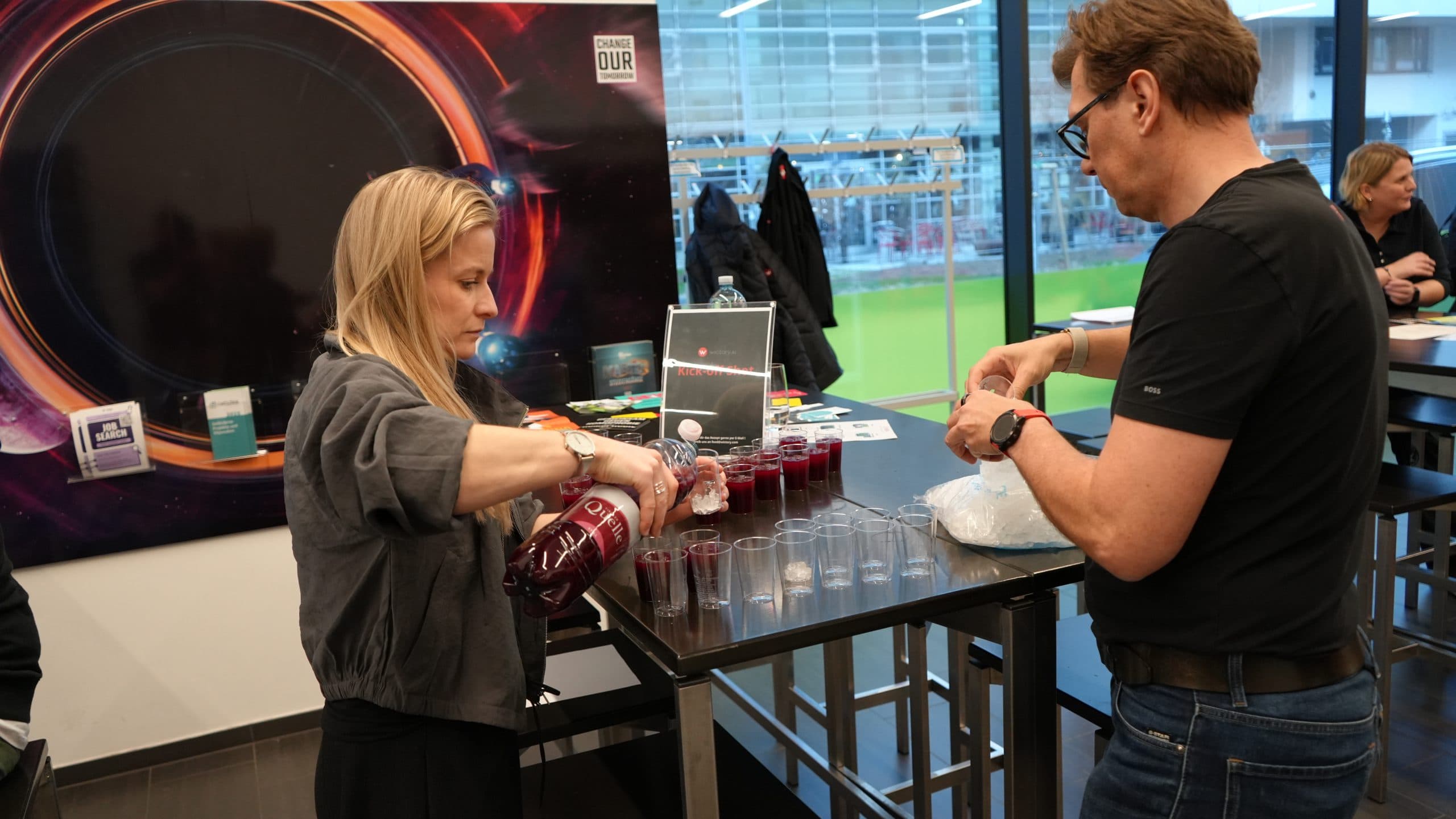 Eine Frau gießt roten Saft in Plastikbecher ein, während ein Mann neben ihr mit einem Beutel Eis hantiert. Sie bereiten die Getränke auf einem Tisch in einem modernen Innenraum vor, in dem sich im Hintergrund weitere Personen befinden.