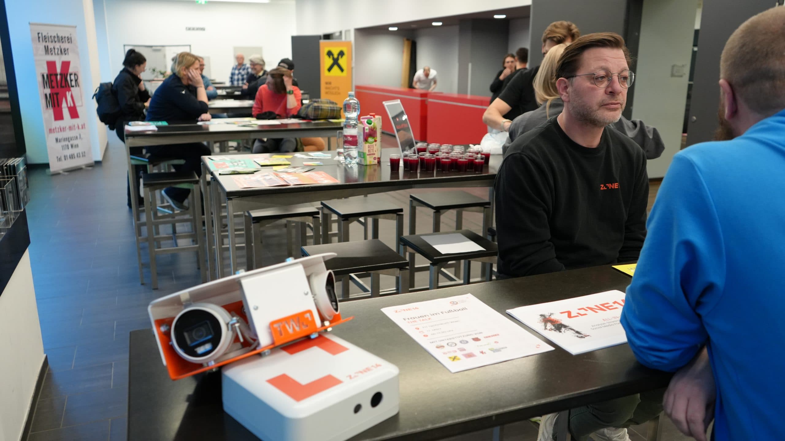 In einem modernen Innenraum sitzen Menschen an Tischen und sind in Gespräche und Aktivitäten vertieft. Auf den Tischen liegen Broschüren und Geräte, und im Hintergrund sind Informationsplakate zu sehen. Die Atmosphäre wirkt locker und organisiert.