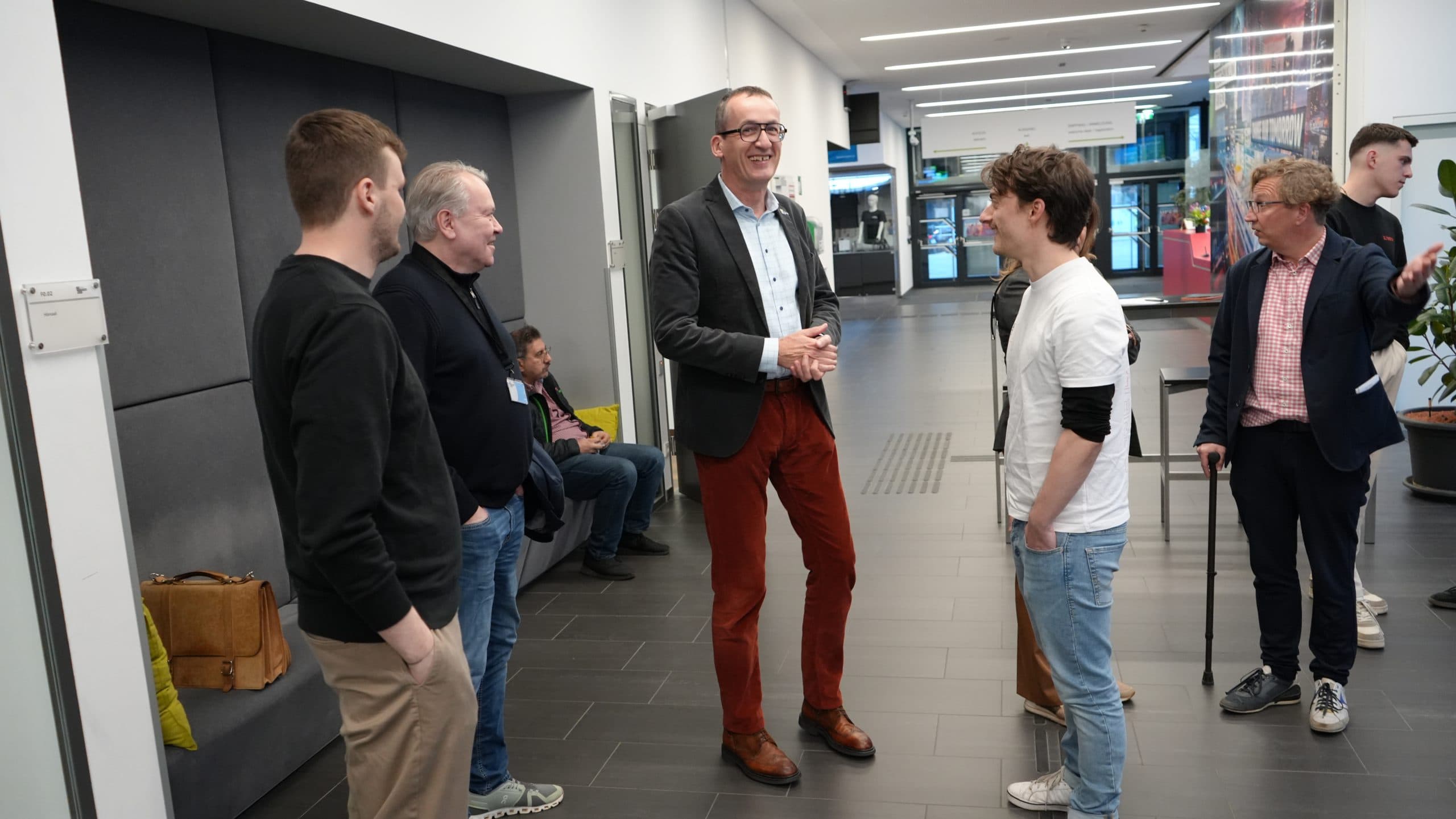 Eine Gruppe von Menschen steht in einem modernen Hausflur und unterhält sich. Ein Mann in Anzug und roter Hose scheint zu der Gruppe zu sprechen, während andere zuhören und lächeln. Einige sind leger gekleidet, und ein paar Leute sitzen im Hintergrund.