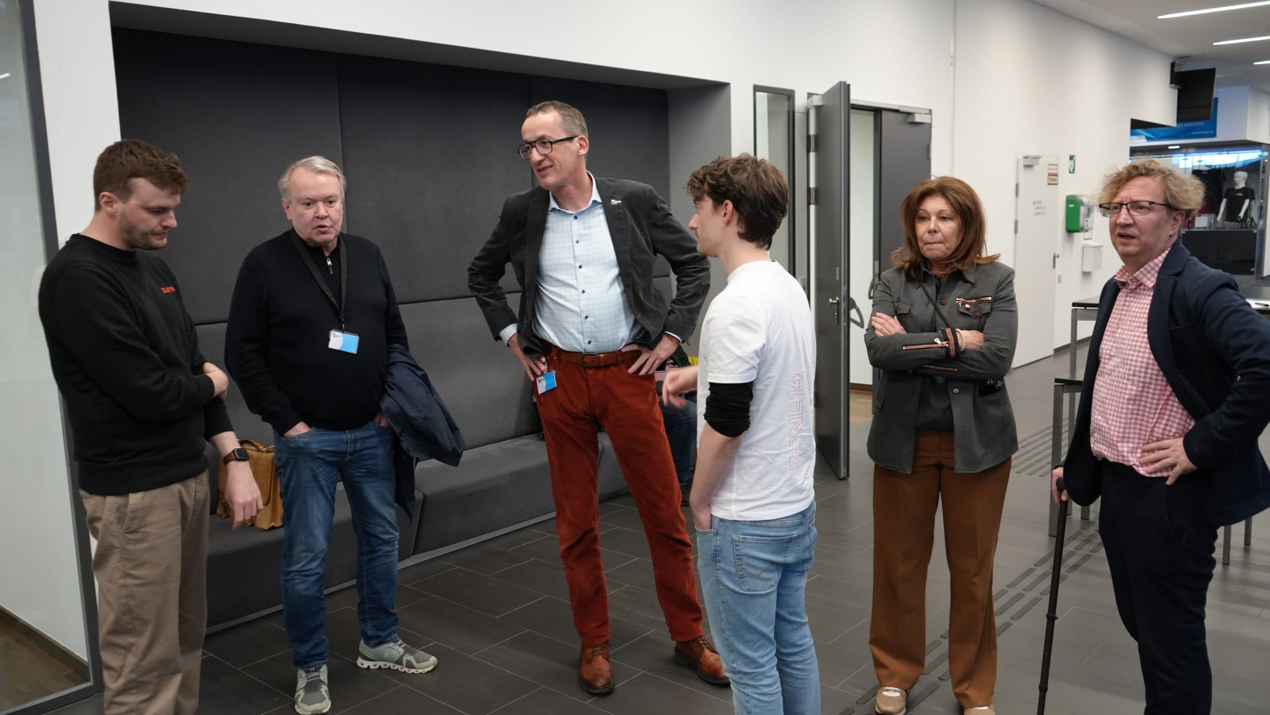 Sechs Personen stehen in einem modernen Hausflur und unterhalten sich. Drei Männer stehen auf der linken Seite, während zwei Männer und eine Frau ihnen auf der rechten Seite gegenüberstehen. Alle sind leger gekleidet und scheinen in ein Gespräch vertieft zu sein.