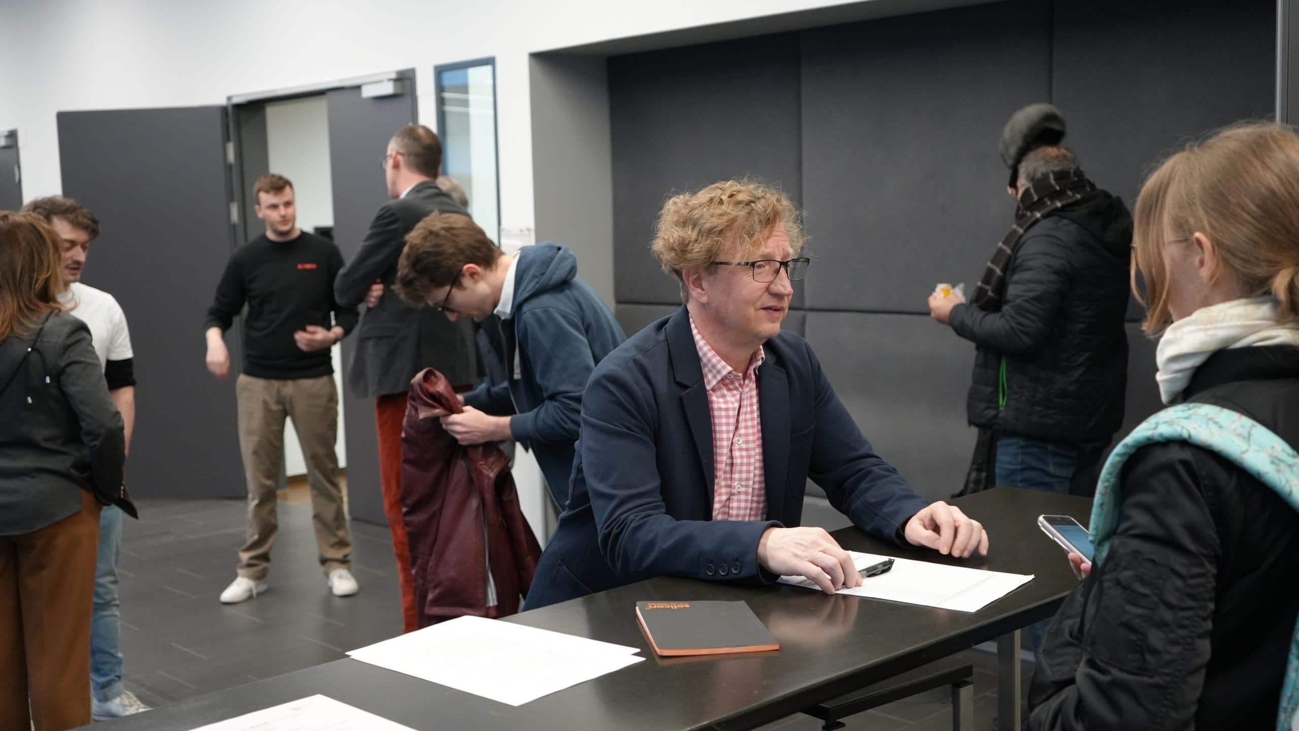 Ein Mann in einem Blazer sitzt an einem Tisch mit Papieren und einem Notizbuch und spricht mit einer Frau, die ein Telefon in der Hand hält. Mehrere andere Personen stehen und sprechen im Hintergrund in einem Flur oder einer Lobby.