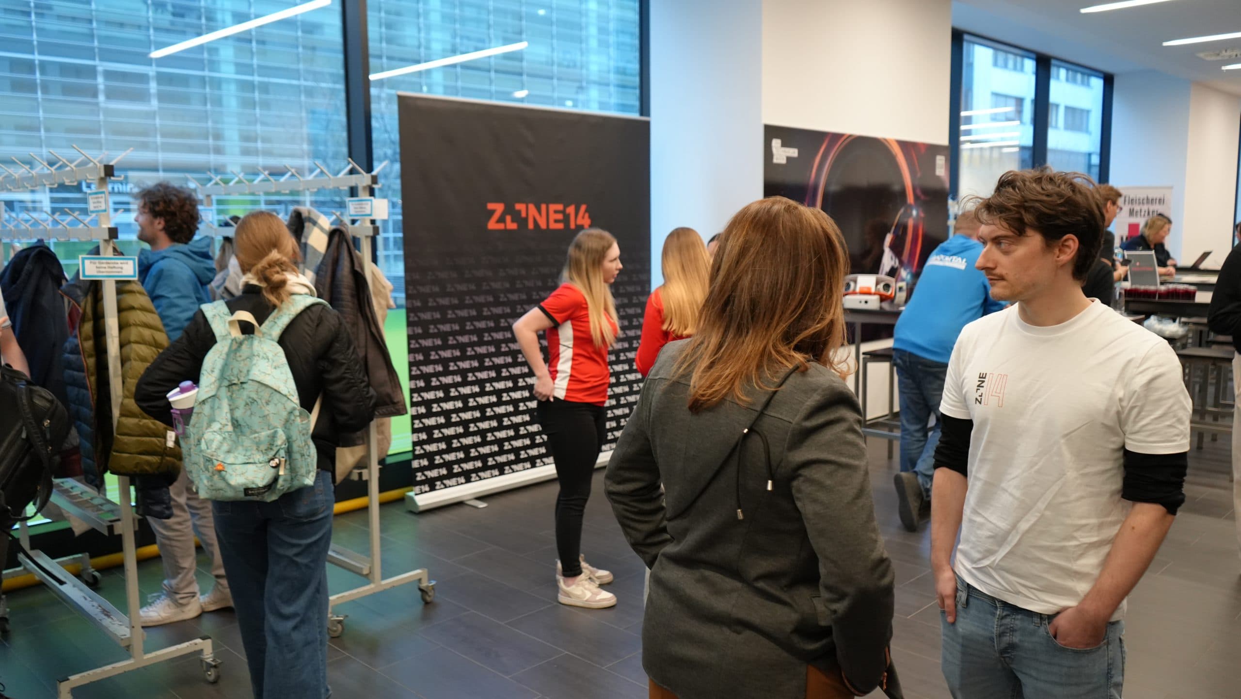 Menschen sind in einem hellen Innenraum mit großen Fenstern versammelt. Einige stehen und unterhalten sich, während andere in der Nähe einer Garderobe stehen. Im Hintergrund ist ein Transparent mit der Aufschrift ZONE14 zu sehen.