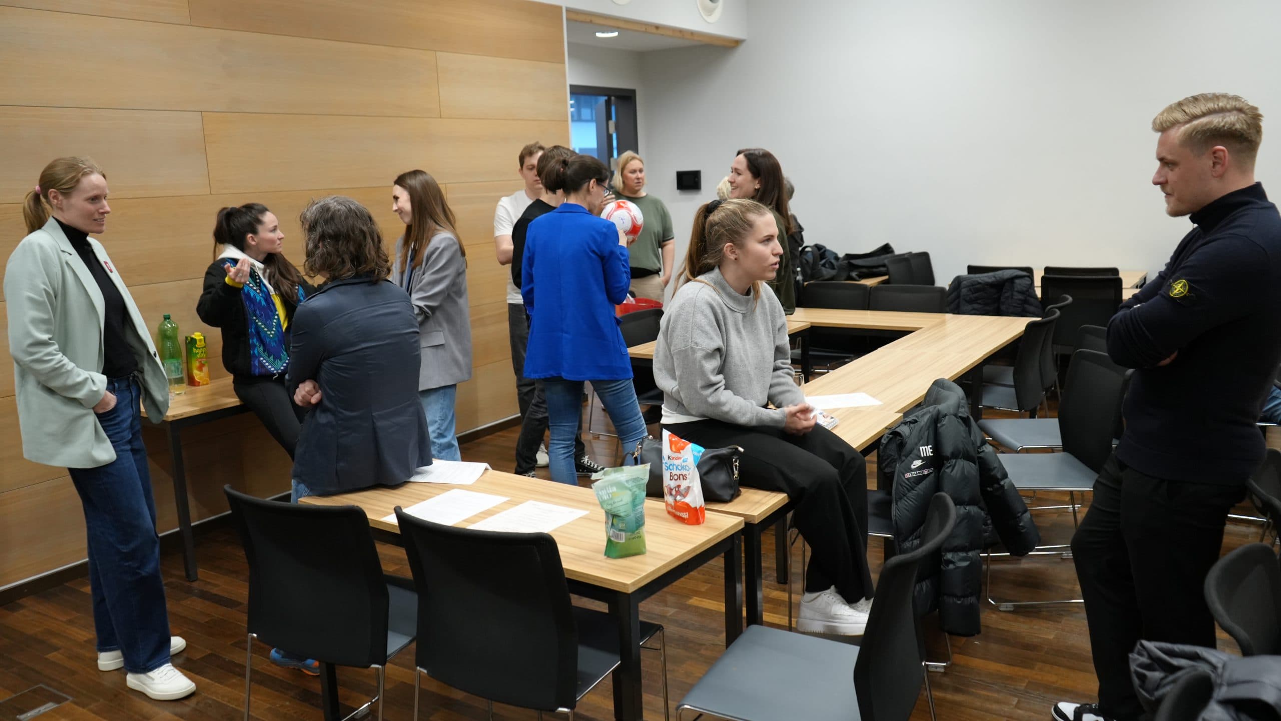 In einem Klassenzimmer oder einem Besprechungsraum steht oder sitzt eine Gruppe von Menschen um Tische herum und unterhält sich. Auf den Tischen und Stühlen liegen Papiere, Flaschen und Jacken. Die Atmosphäre scheint informell und entspannt zu sein.
