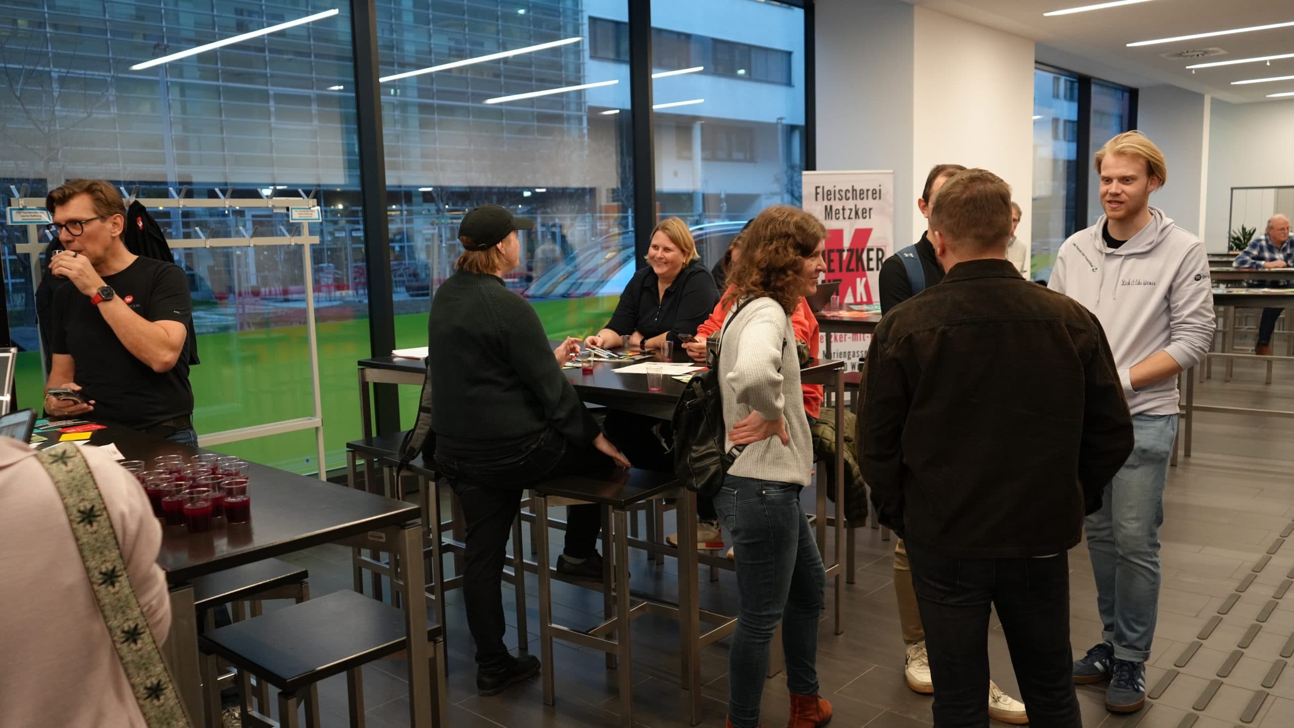 Eine Gruppe von Menschen sitzt an Stehtischen in einem modernen Innenraum mit großen Fenstern zusammen. Einige sitzen, andere stehen und unterhalten sich, auf den Tischen stehen Getränke und Snacks.