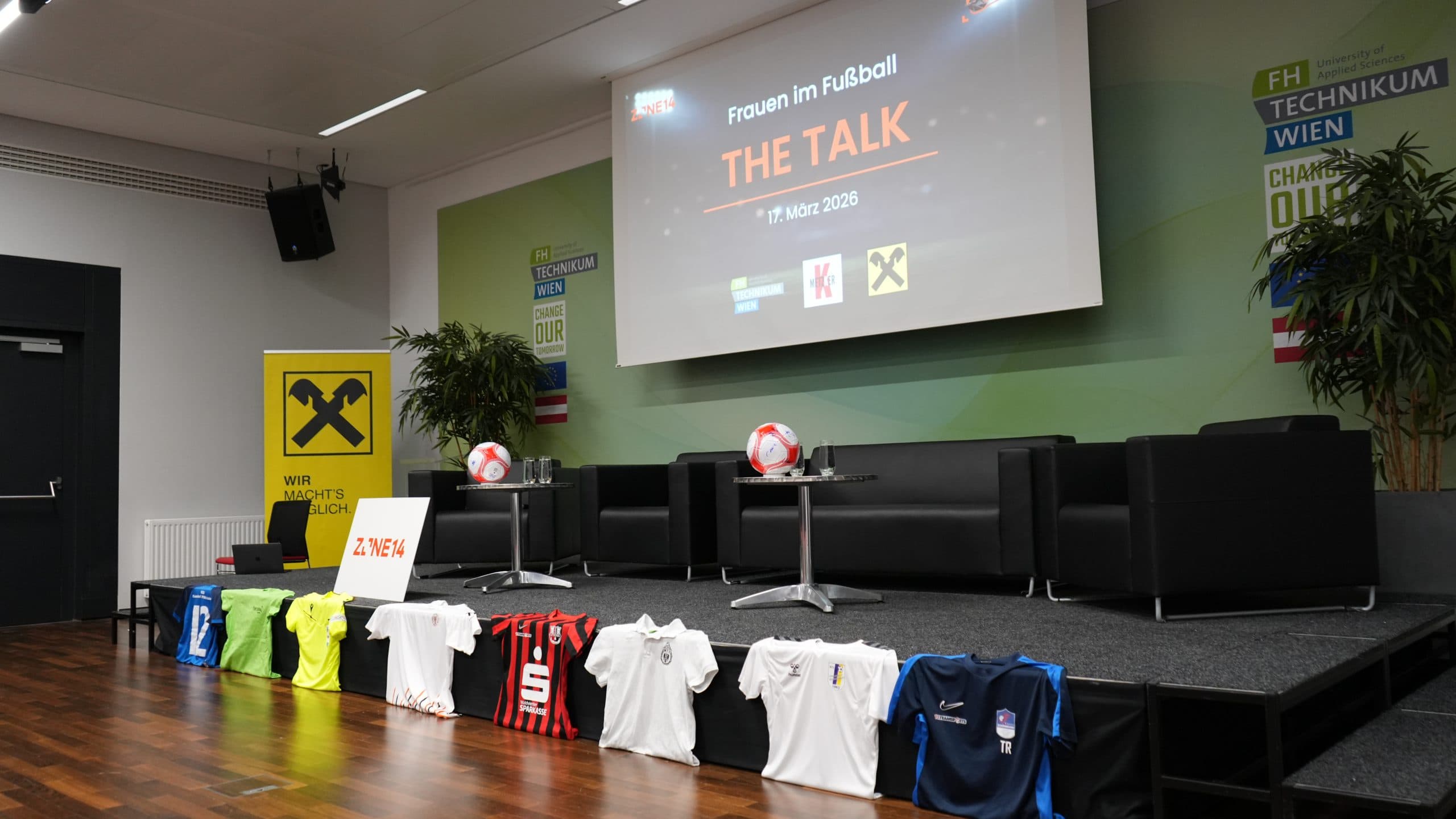 Ein Bühnenbild für eine Fußball-Talk-Veranstaltung mit Stühlen, Tischen, zwei Fußbällen und Trikots verschiedener Mannschaften im Vordergrund. Auf einer Leinwand im Hintergrund ist zu lesen: 'Frauen im Fußball THE TALK, 1. März 2026'.