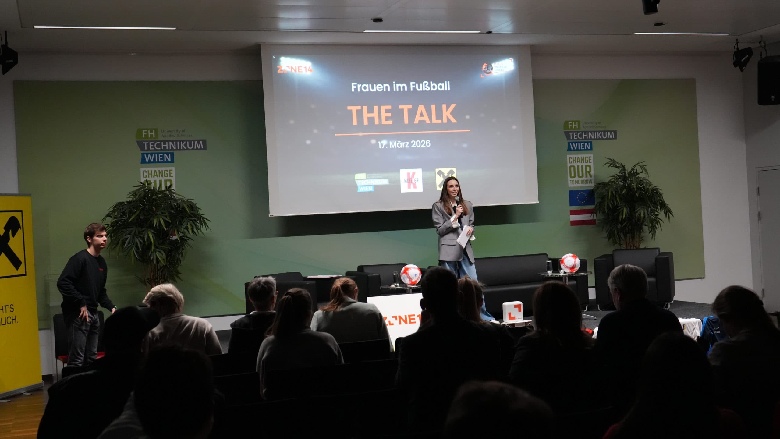 Eine Frau steht auf der Bühne und spricht bei einer Veranstaltung von Frauen im Fußball in ein Mikrofon. Hinter ihr befindet sich eine Präsentationsfolie mit der Aufschrift 'THE TALK'. Die Zuhörer sitzen in einem modernen Hörsaal mit Blick auf die Bühne.
