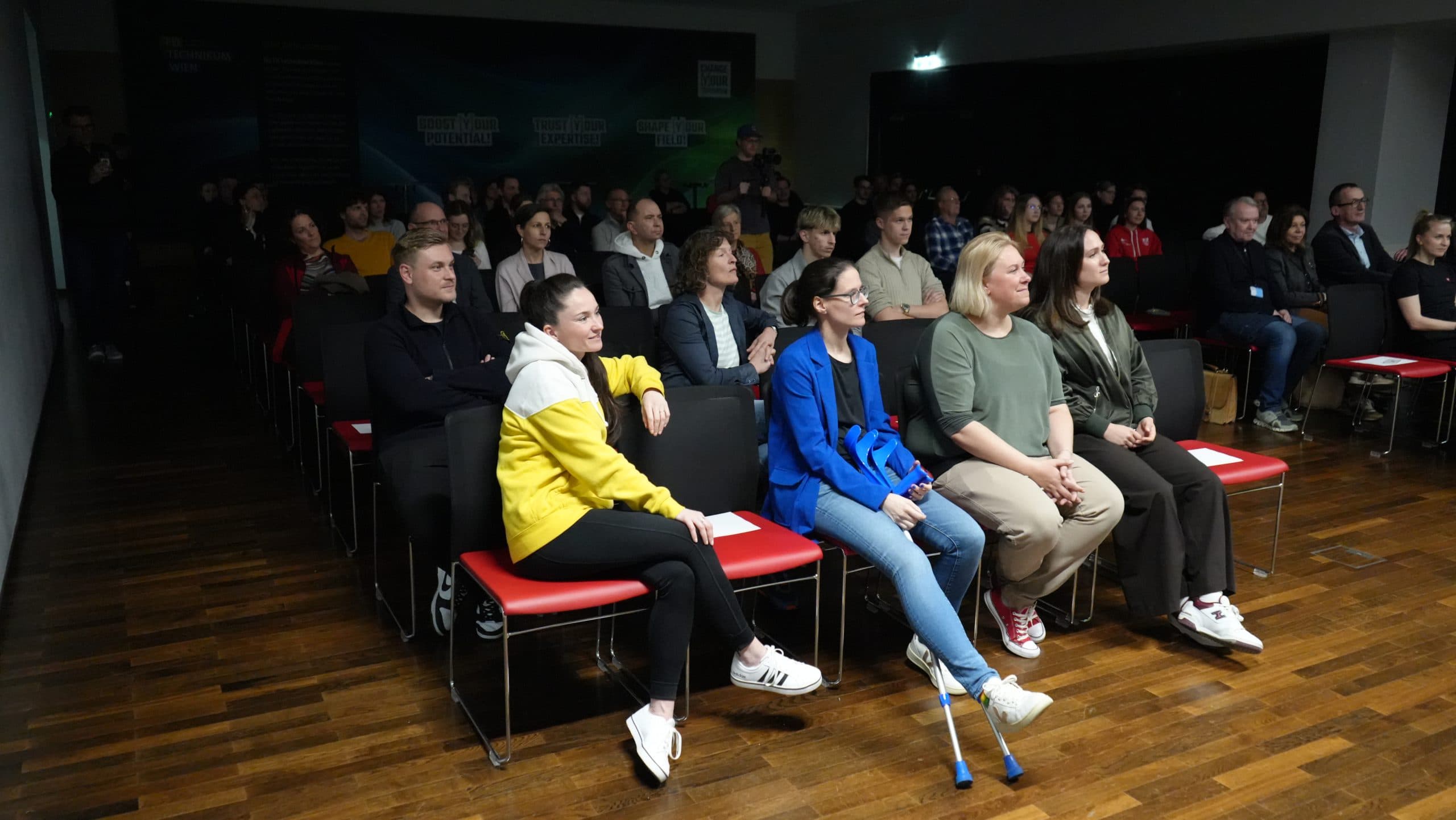 Die Zuschauer sitzen auf Stühlen in einem schwach beleuchteten Raum und beobachten aufmerksam etwas aus dem Off. Die Gruppe ist bunt gemischt, mit Männern und Frauen unterschiedlichen Alters; eine Frau in der ersten Reihe benutzt eine Krücke.
