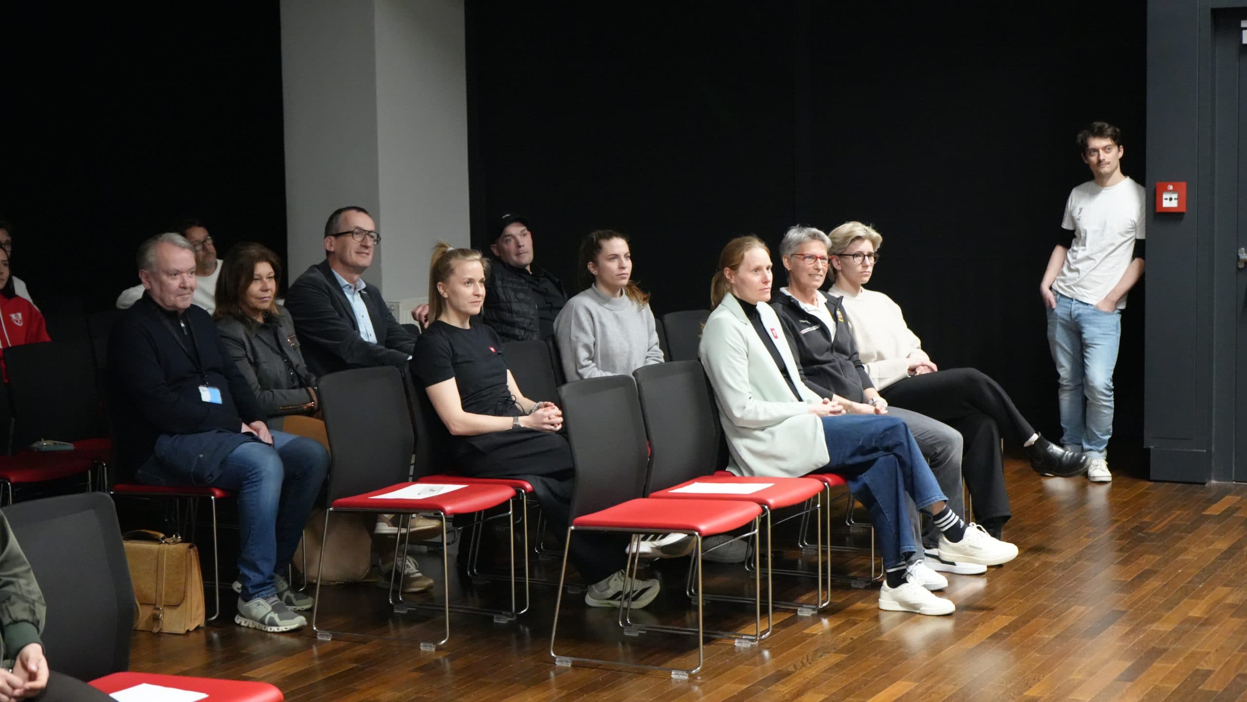 Eine Gruppe von Personen sitzt in Stuhlreihen in einem Raum mit schwarzen Wänden und hört aufmerksam zu. Eine Person steht in der Nähe der Tür auf der rechten Seite und lehnt sich gegen die Wand. Der Boden ist aus Holz, und einige Stühle sind leer.