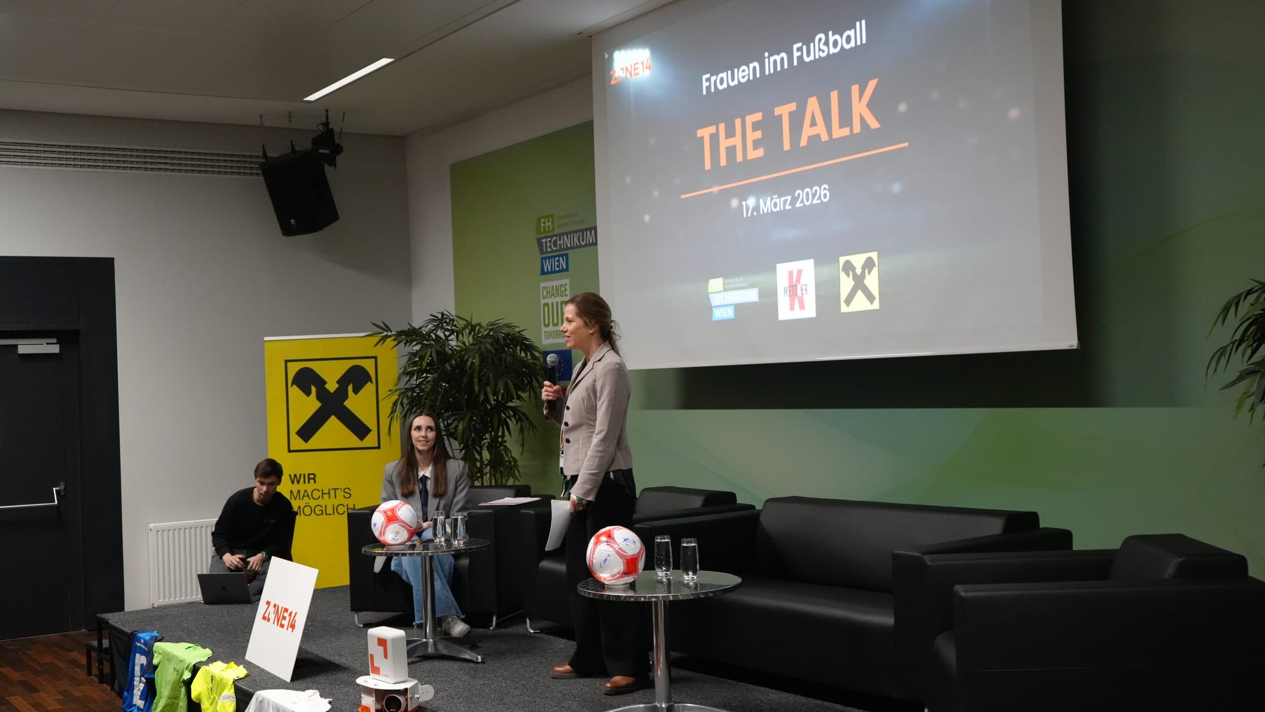 Zwei Frauen stehen auf der Bühne einer Veranstaltung namens 'Frauen im Fußball - THE TALK'. Die eine steht und spricht in ein Mikrofon, die andere sitzt neben ihr. Im Raum sind Gegenstände und Banner zum Thema Fußball zu sehen.