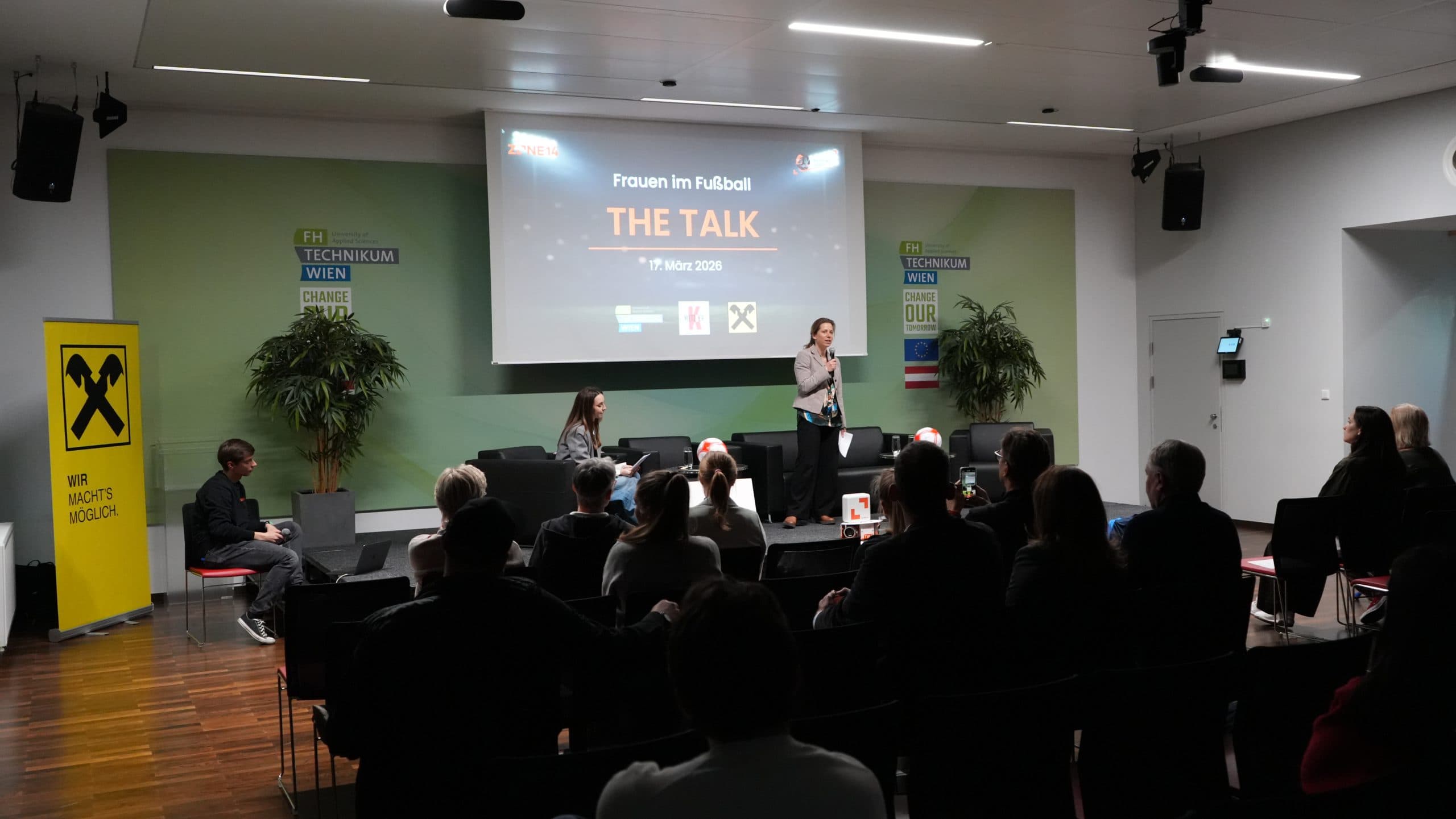 Eine Frau steht auf der Bühne und spricht zu einem Publikum in einem modernen Hörsaal. Auf einer Leinwand hinter ihr steht 'Frauen im Fußball: THE TALK, 7. März 2023'. Die Zuhörer sitzen und hören aufmerksam zu.